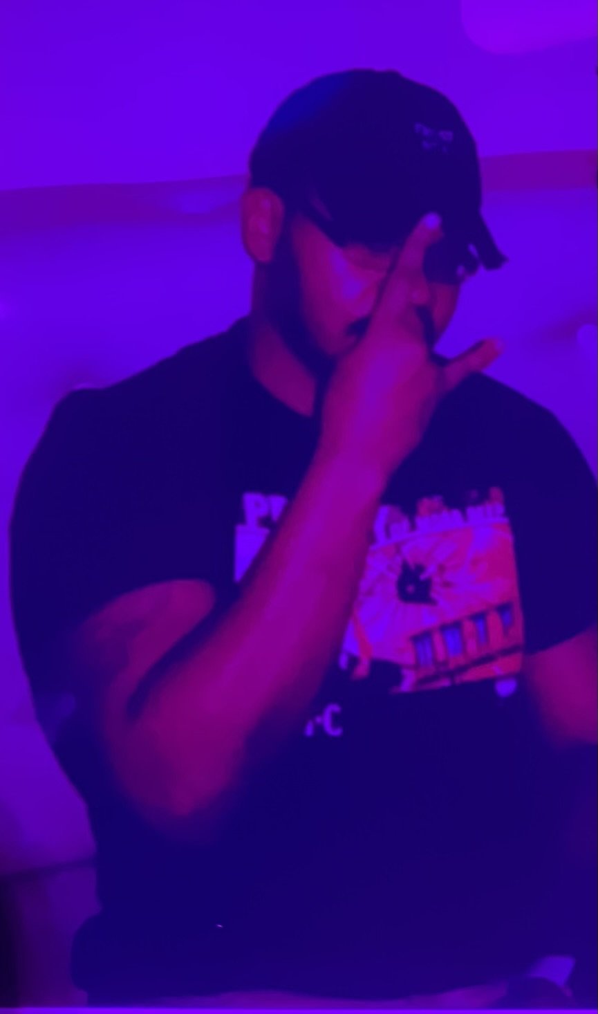 Frandy Scenario with glasses wearing a black t-shirt, Black hat, is posing with his hand covering part of their face in a room lit with purple and pink neon lights.