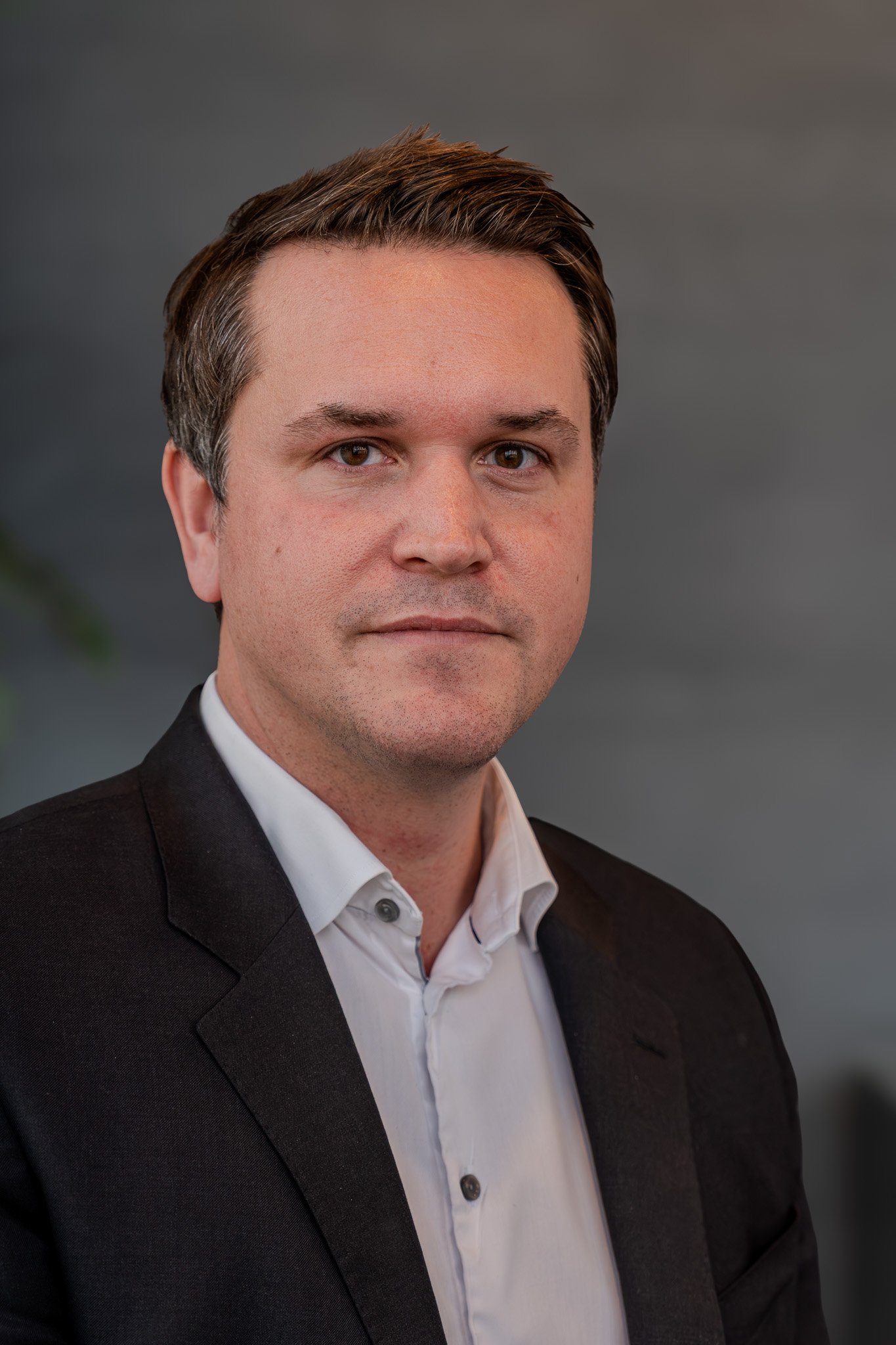 Porträtt av en man i mörk kostym och vit skjorta, med ljus hud och kort, mörkt hår, mot en grå bakgrund.