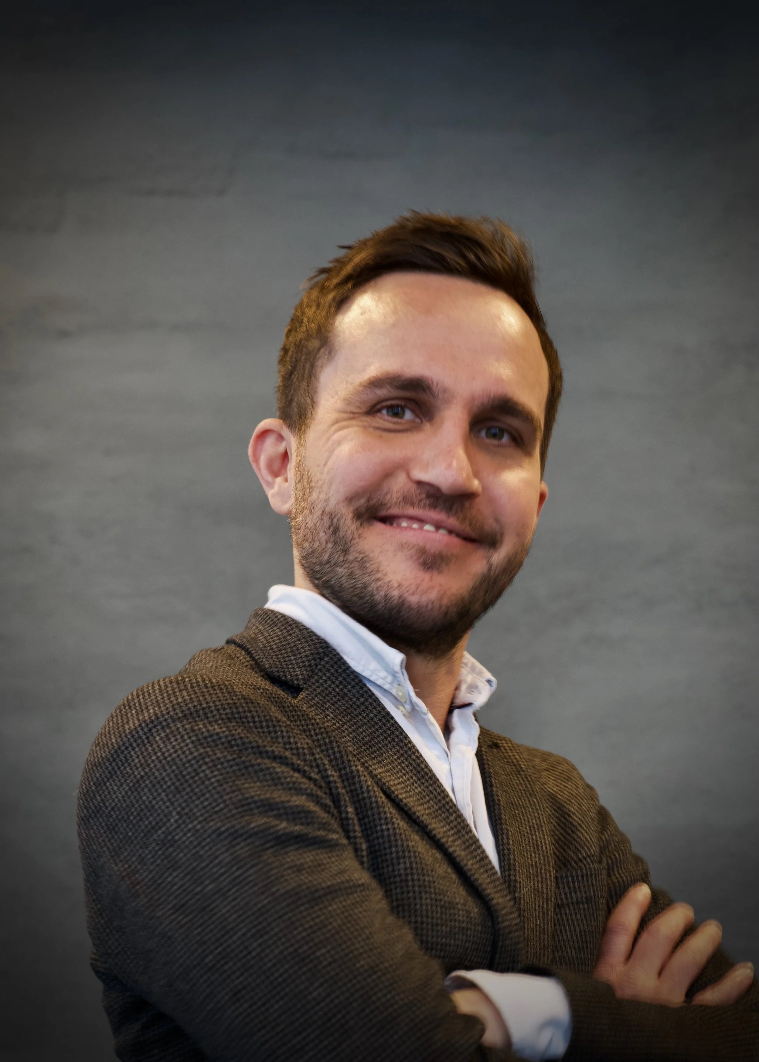 Portrett av en smilende mann i dress, med armene krysset, mot en mørk bakgrunn.