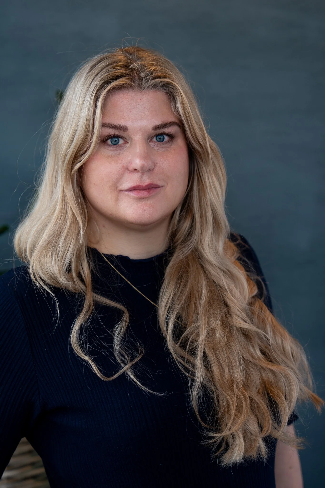 Portrett av en kvinne med lang, blond, bølget hår, iført svart genser, mot en grå bakgrunn.