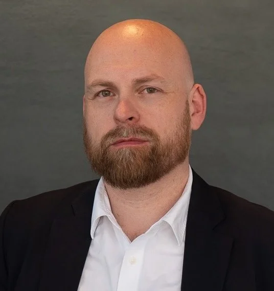 Portrett av en mann med skjegg, som har på seg en hvit skjorte og svart blazer, mot en grå bakgrunn.