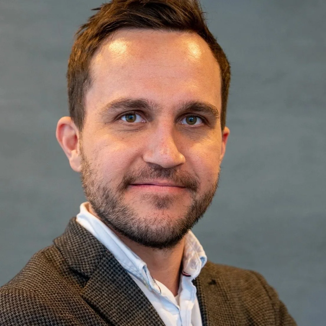 Portrett av en mann med kort brunt hår og skjegg, iført en hvit skjorte og brun blazer, poserer mot en grå bakgrunn.