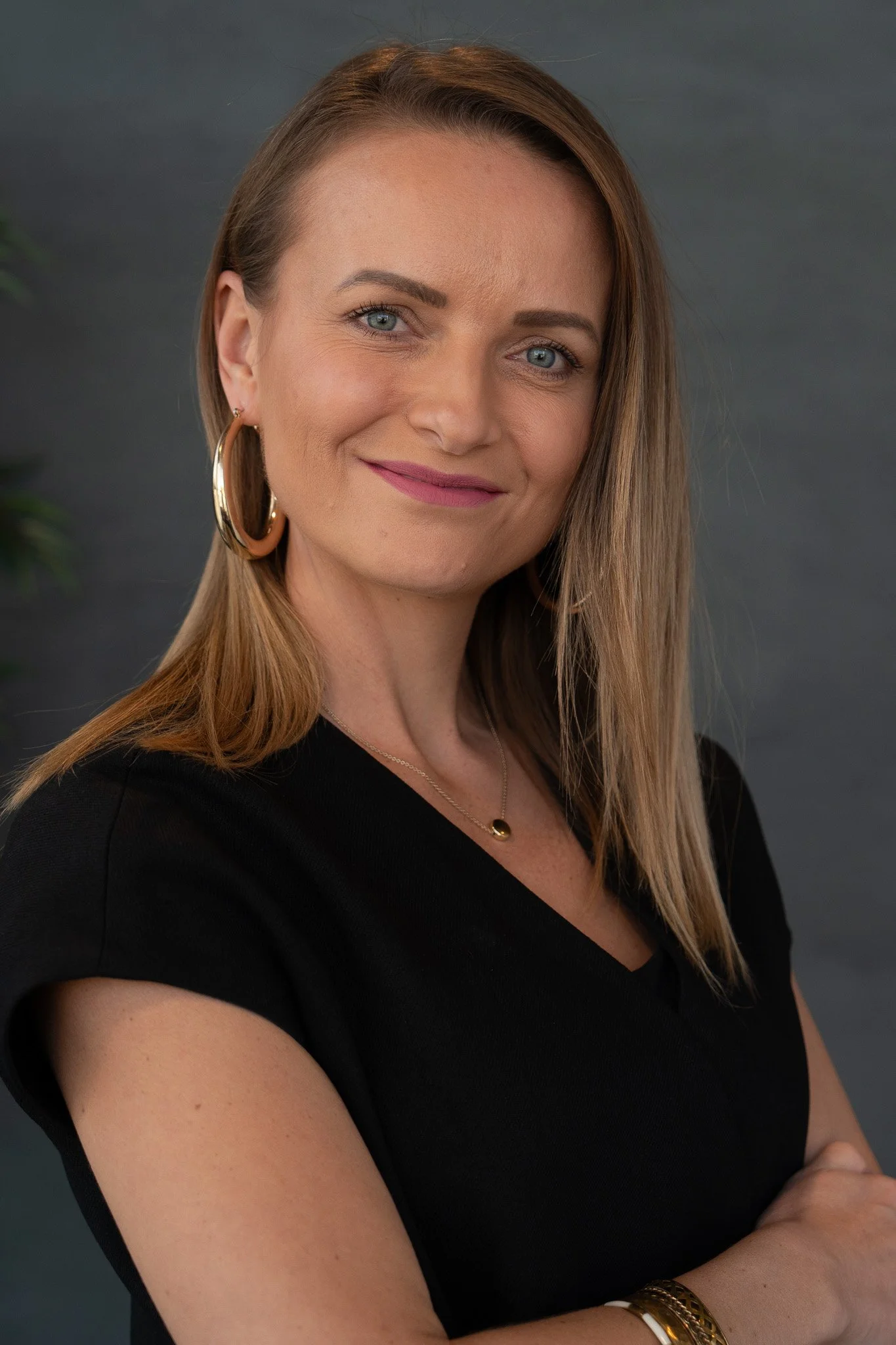 Portrett av en kvinne med skulderlangt hår, iført svart topp, gull ørefigurer og smykker, smilende mot kameraet.