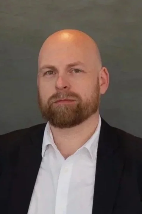 Portrett av en mann med skjegg, som har på seg en hvit skjorte og svart blazer, mot en grå bakgrunn.