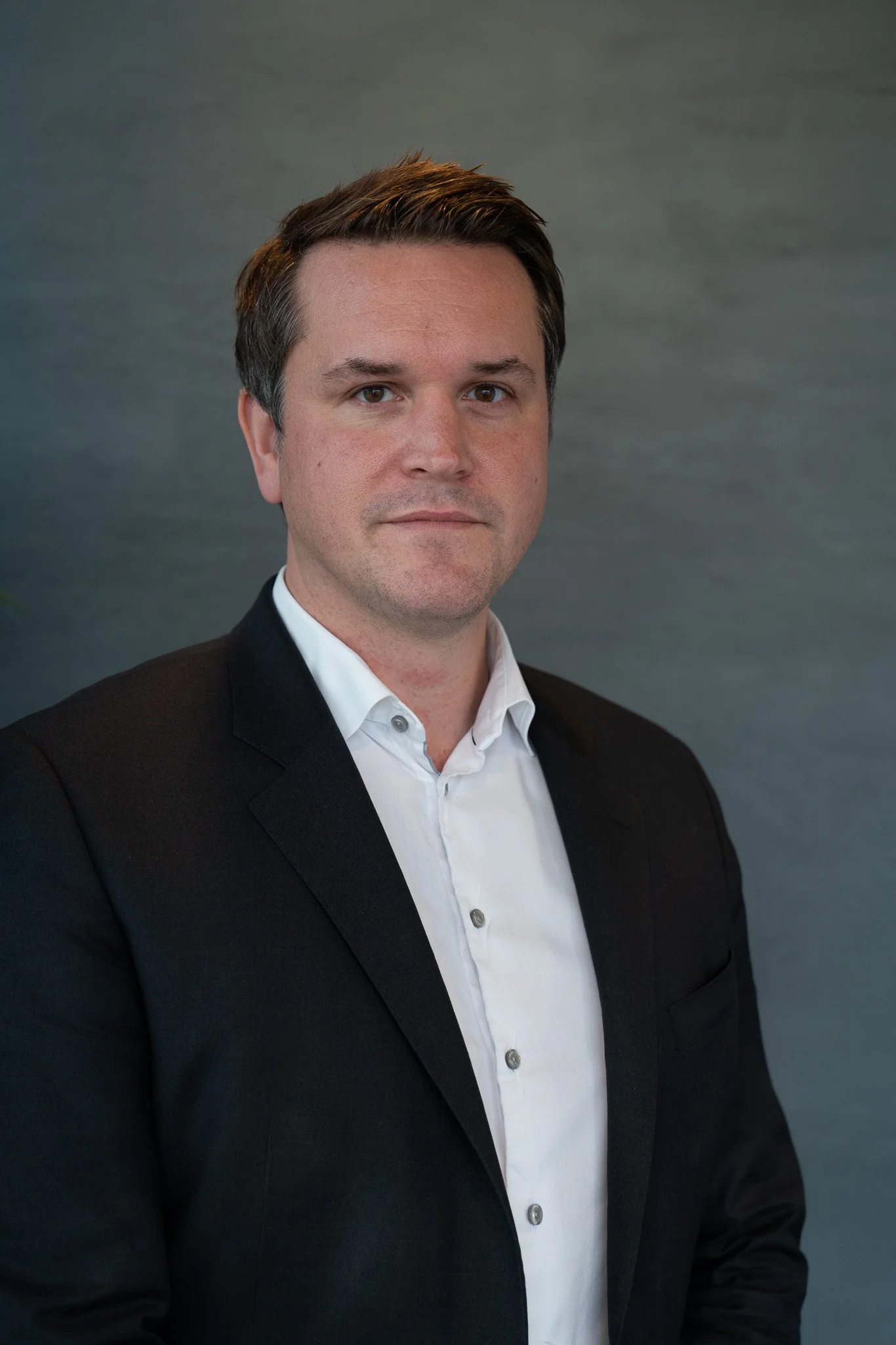 Profesjonell portrett av en mann i svart dress og hvit skjorte mot en grå bakgrunn.