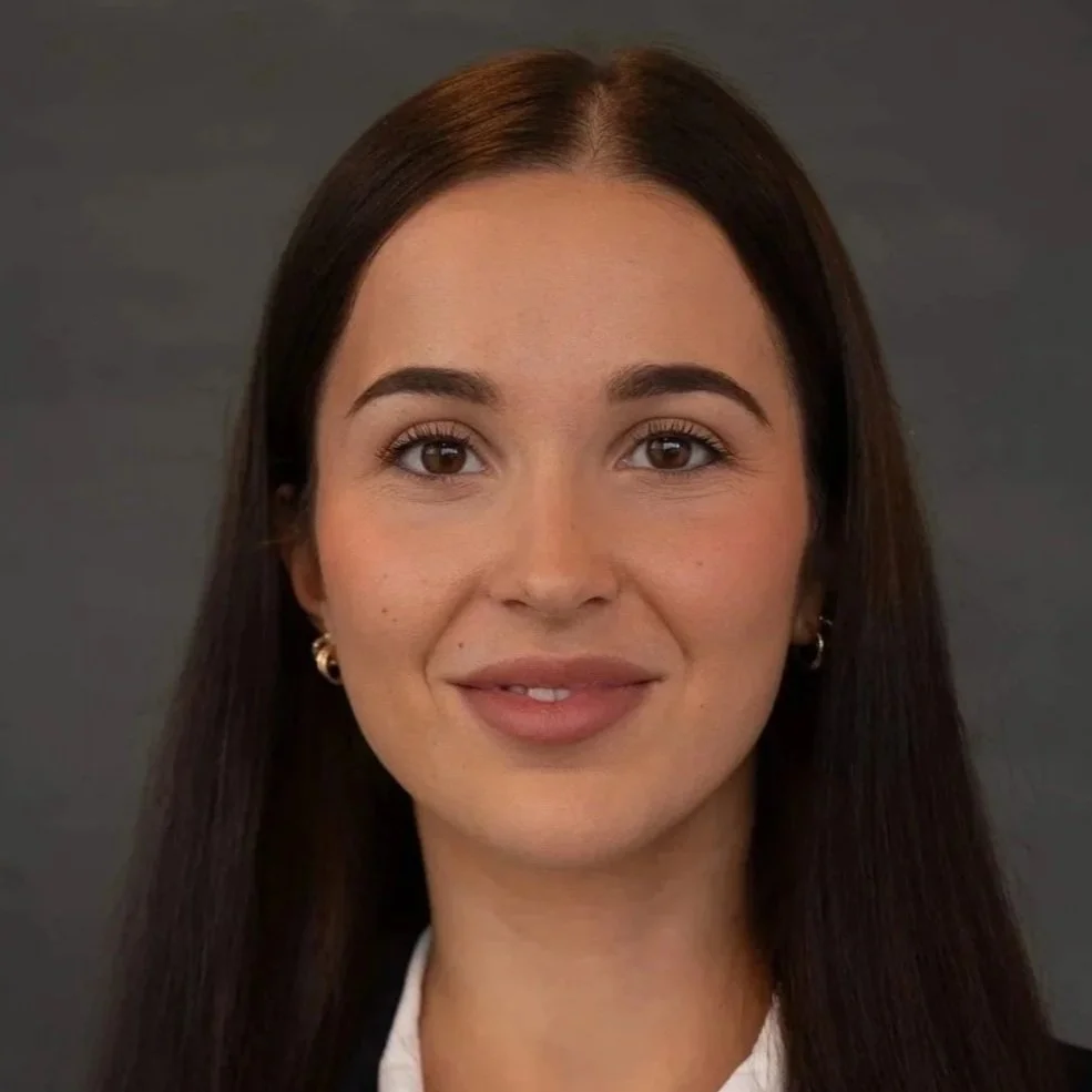 Portrett av en ung kvinne med mørkt, langt hår og smilende uttrykk, iført hvit skjorte og små øreringer mot en grå bakgrunn.