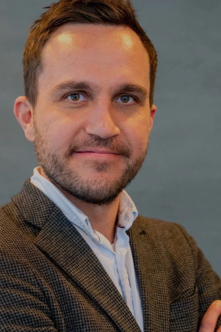Portrett av en mann med kort brunt hår og skjegg, iført en hvit skjorte og brun blazer, poserer mot en grå bakgrunn.
