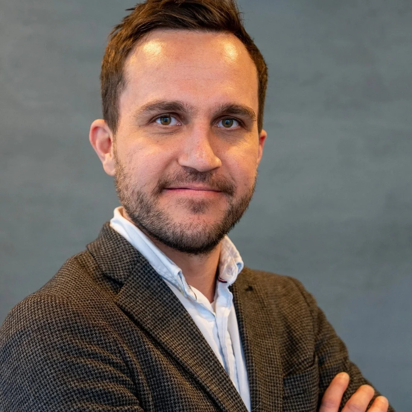 Portrett av en mann med mørkt hår og skjegg, iført en brun jakke og hvit skjorte, mot en grå bakgrunn.