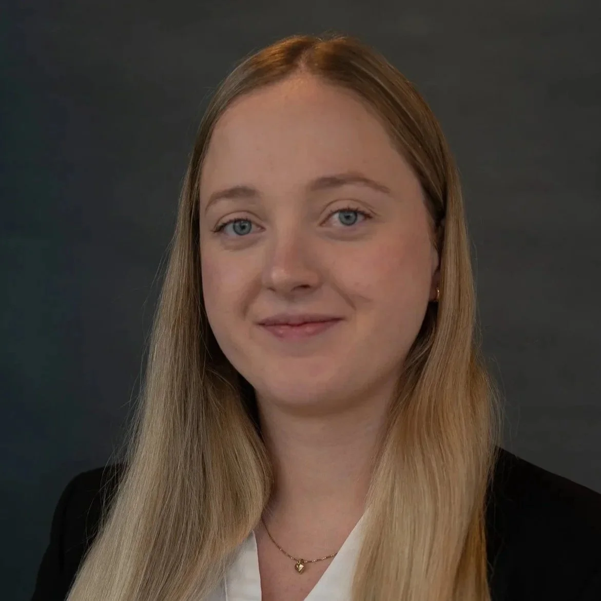 Portrett av en kvinne med lang blond hår, blå øyne, smilende, iført svart jakke og hvit skjorte, mot en mørk bakgrunn.