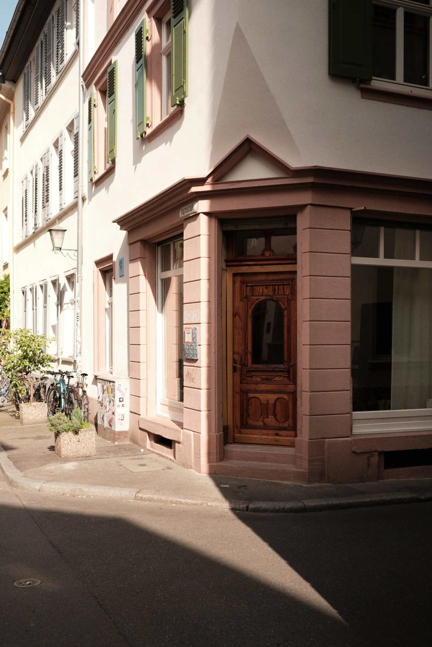 Ecke eines Gebäudes mit Holztür, rosafarbener Steinfassade und grünen Fensterläden. Fahrräder stehen auf dem Gehweg vor dem Fotostudio in Heidelberg.