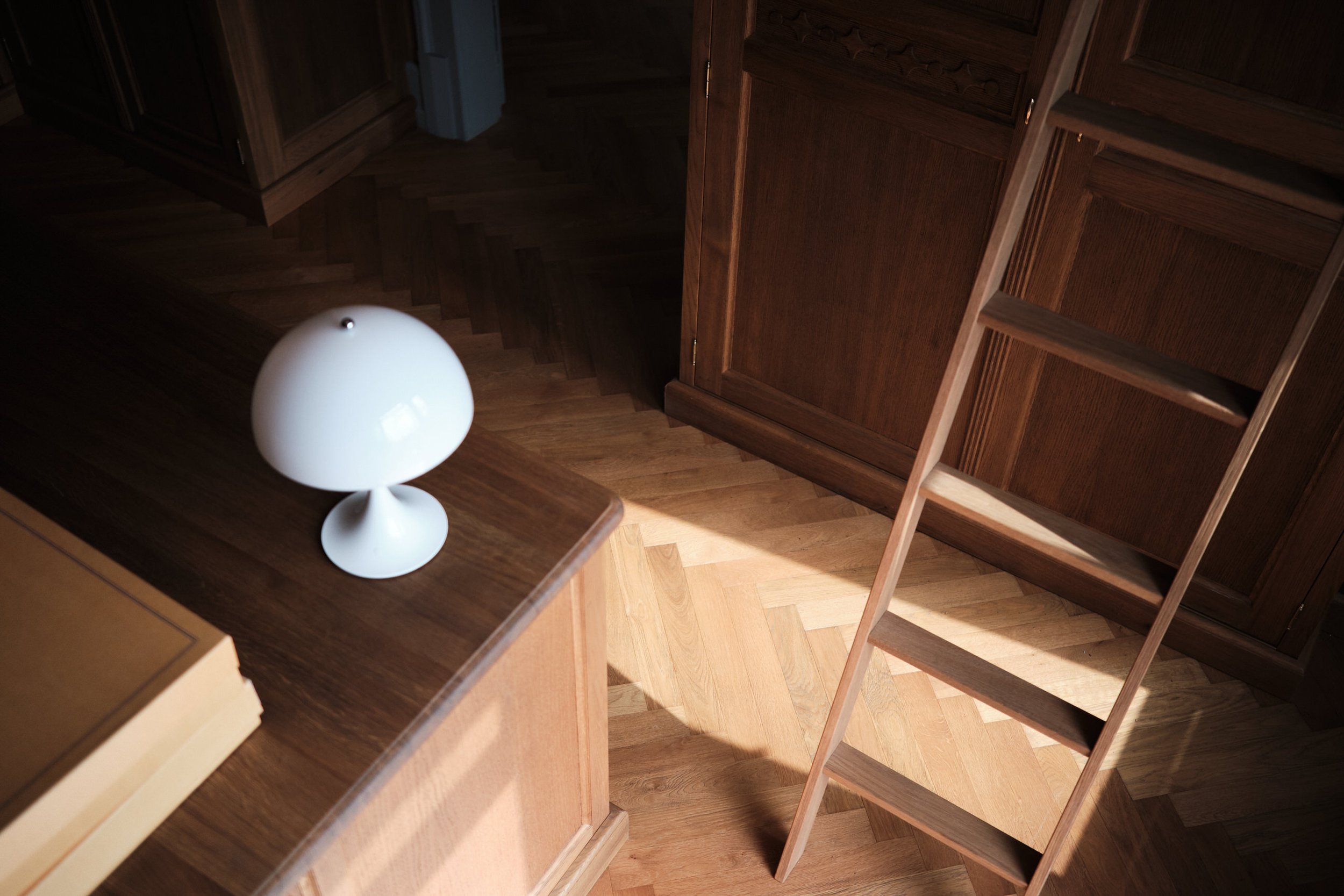 Weiße, moderne Tischleuchte auf einer Holzarbeitsfläche neben einer Holzleiter, die an dunkle Holzschränke in einem Küchen oder Arbeitsbereich lehnt, fotografiert im Rahmen einer geschmackvollen Architekturfotografie.
