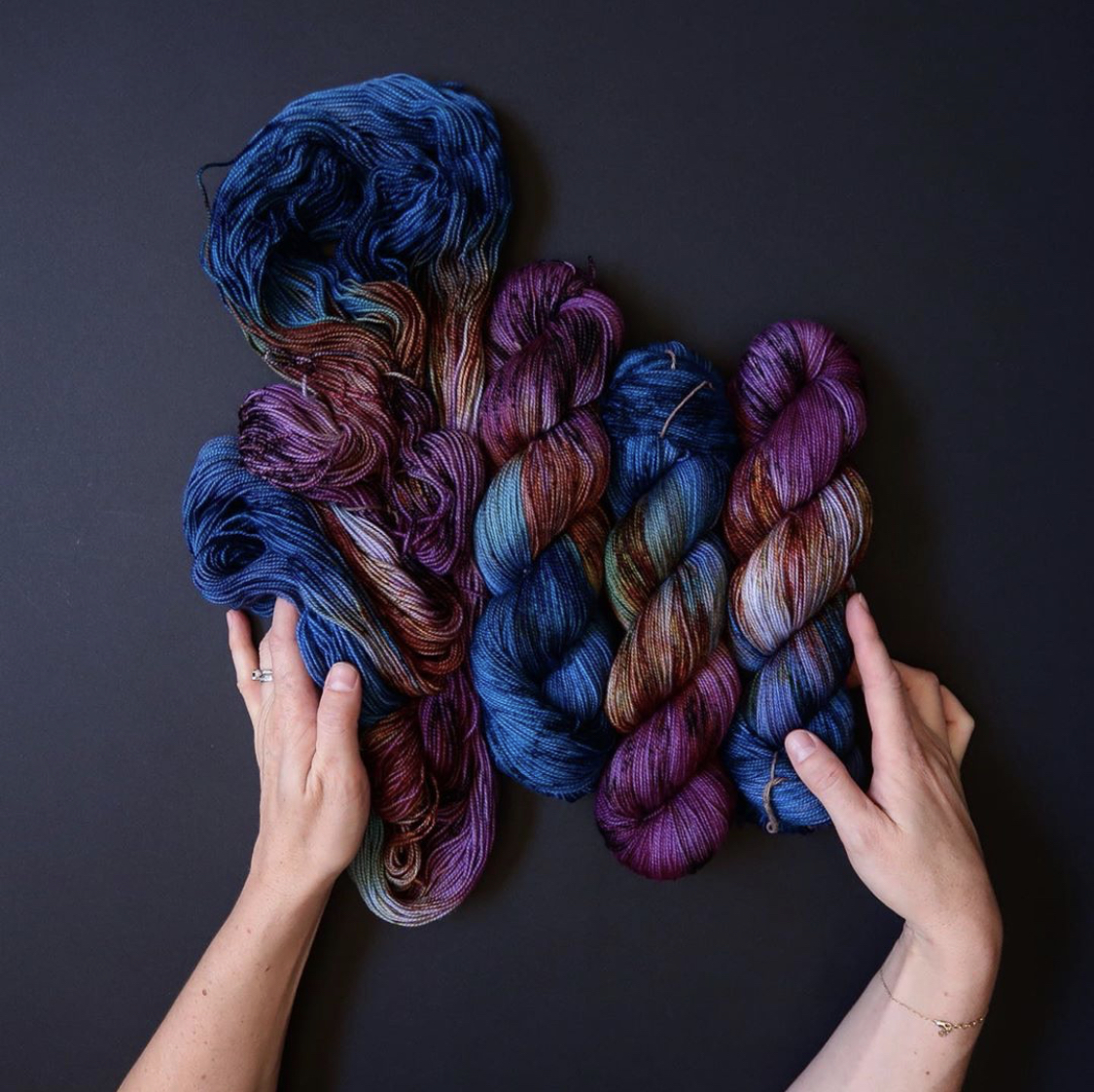 Tayler’s hands embrace four hanks of blue & purple yarn, one hank unskeined and flowing; Photo by Tayler Earl of Fiber for the People®