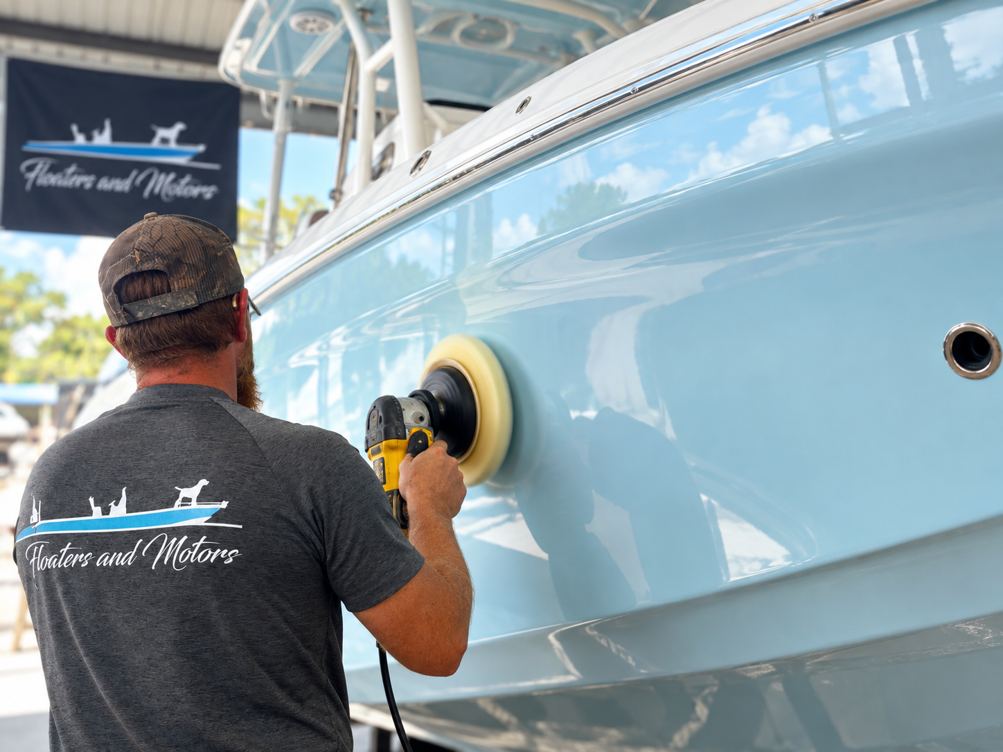 A man polishing the hull of a boat with a power buffer, wearing a gray t-shirt with 'Floaters and Motors' logo on the back, in an outdoor boatyard with blue sky and trees in the background.