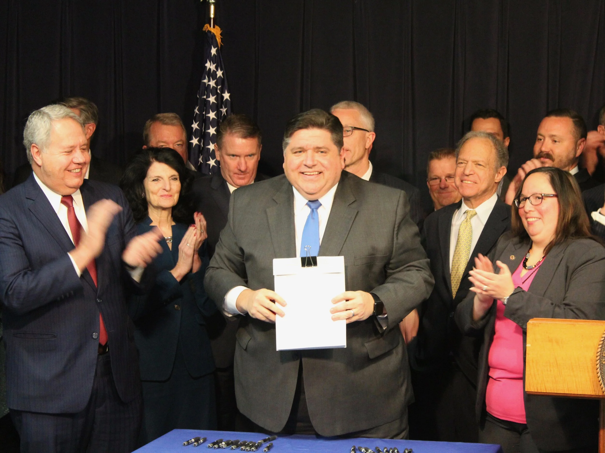 Flanked by lead sponsors Rep. Jay Hoffman and Sen. Cristina Castro and backed by members of the state pension task force and police and firefighter union heads, Gov. Pritzker brandishes the signed bill consolidating 649 local funds. (One Illinois/Te…