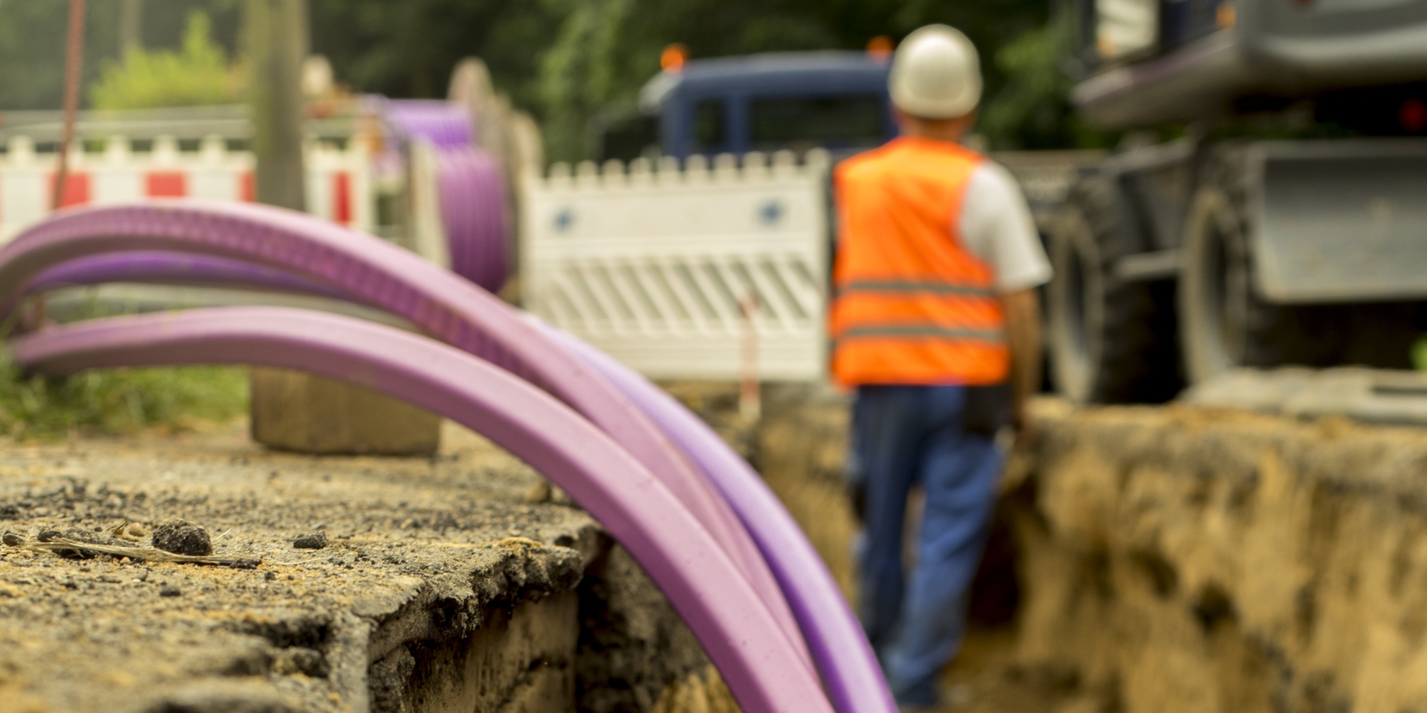 Image: A construction worker stands in a waist-high ditch. Purple cables are in the foreground. 