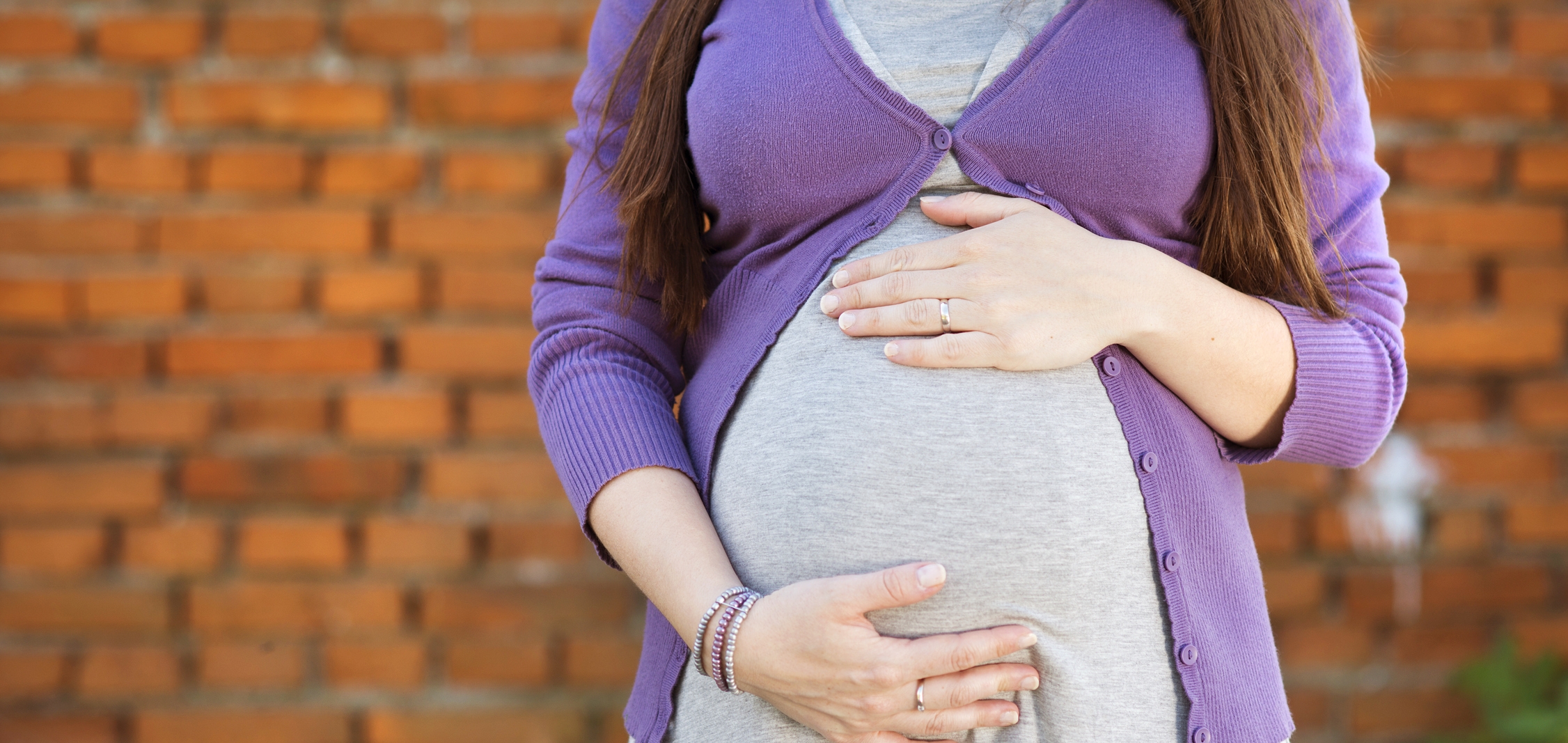 Image: A woman in a gray dress stands in front of a brick wall. She is holding her pregnant stomach. 