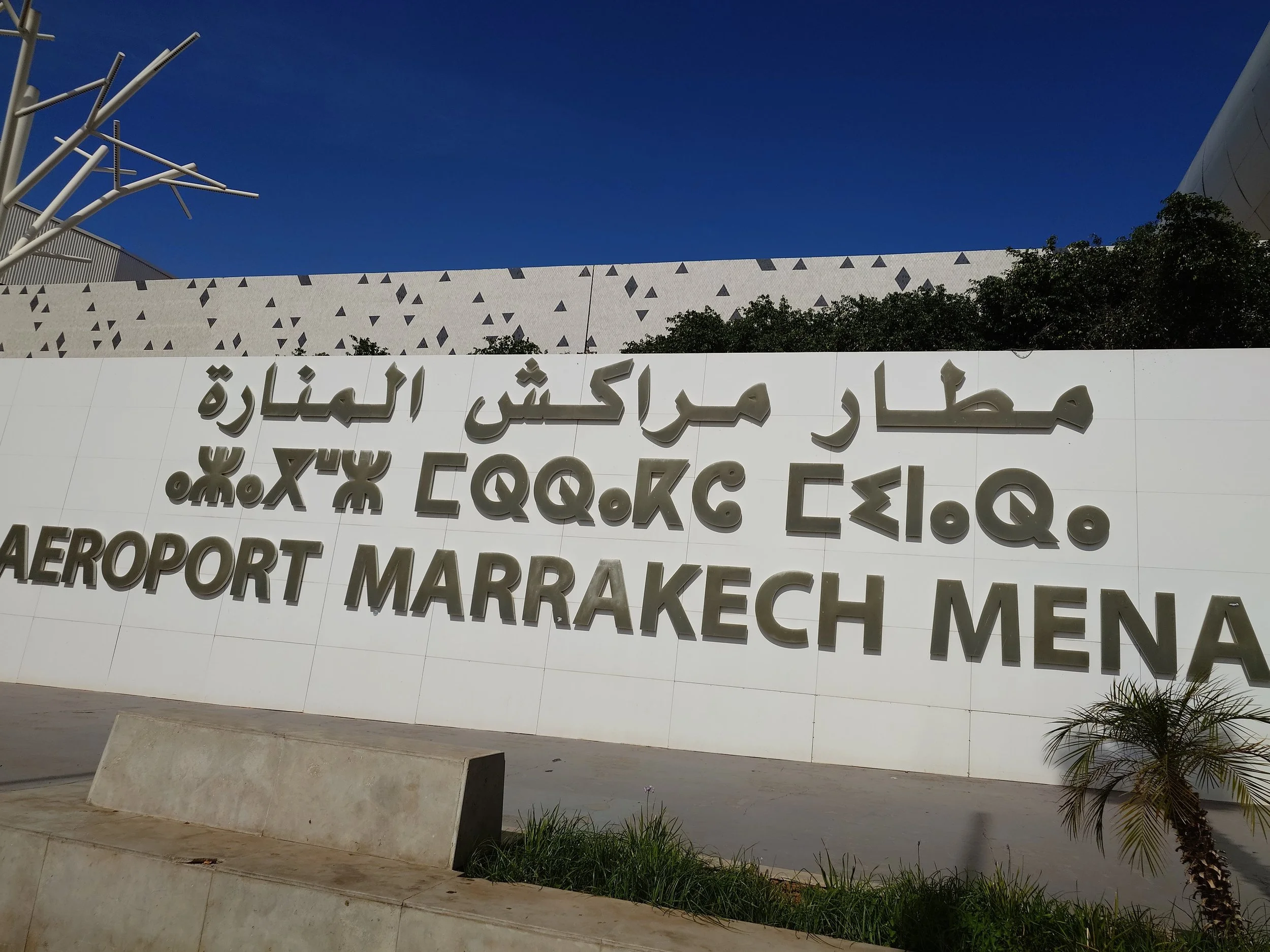 Sign at Marrakech Menara Airport with Arabic, French, and English text on a white background, under a clear sky.