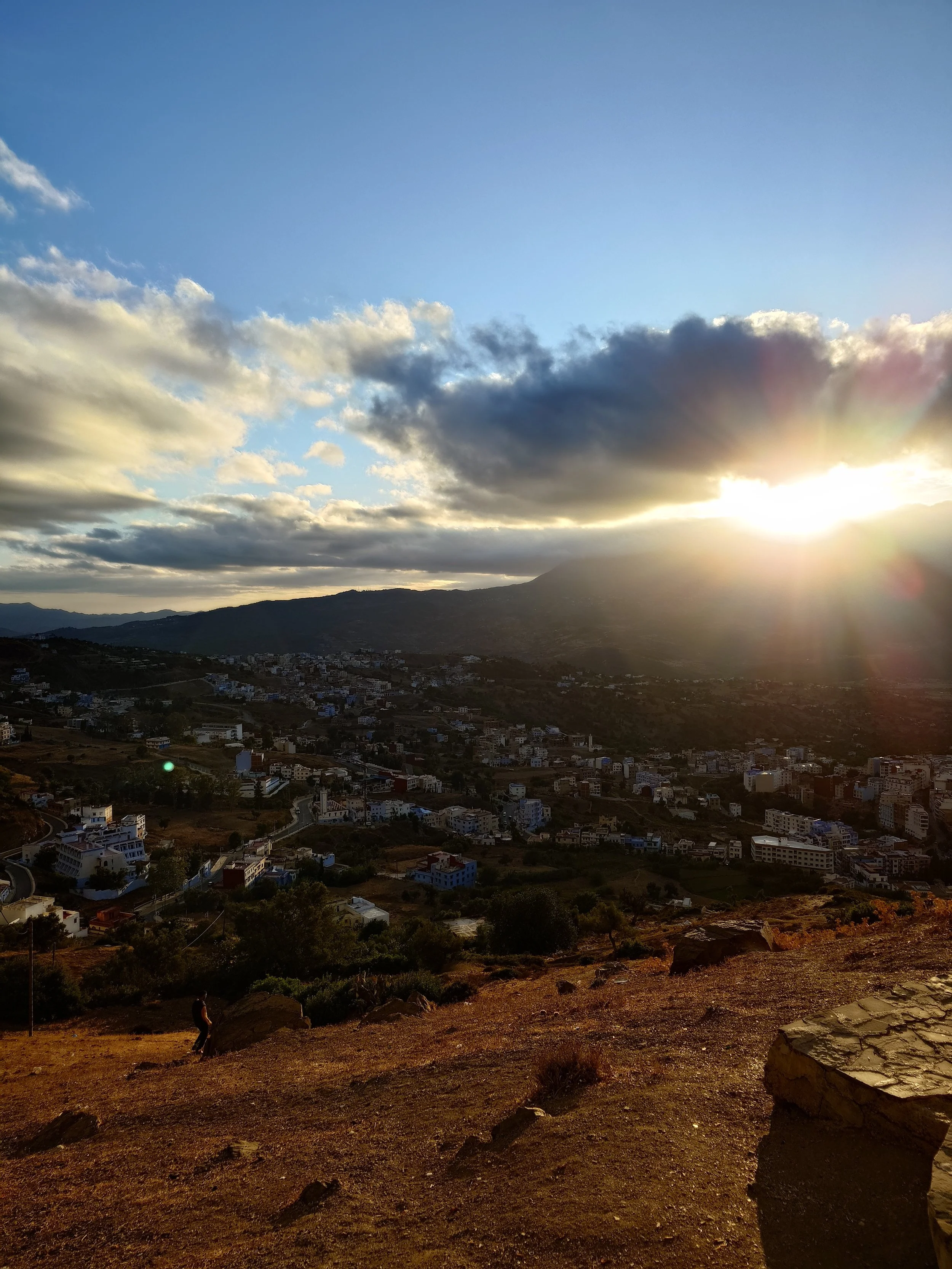 Sunset over a town with mountains in the background, cloudy sky, rocky foreground, and a person walking in the lower-left corner.