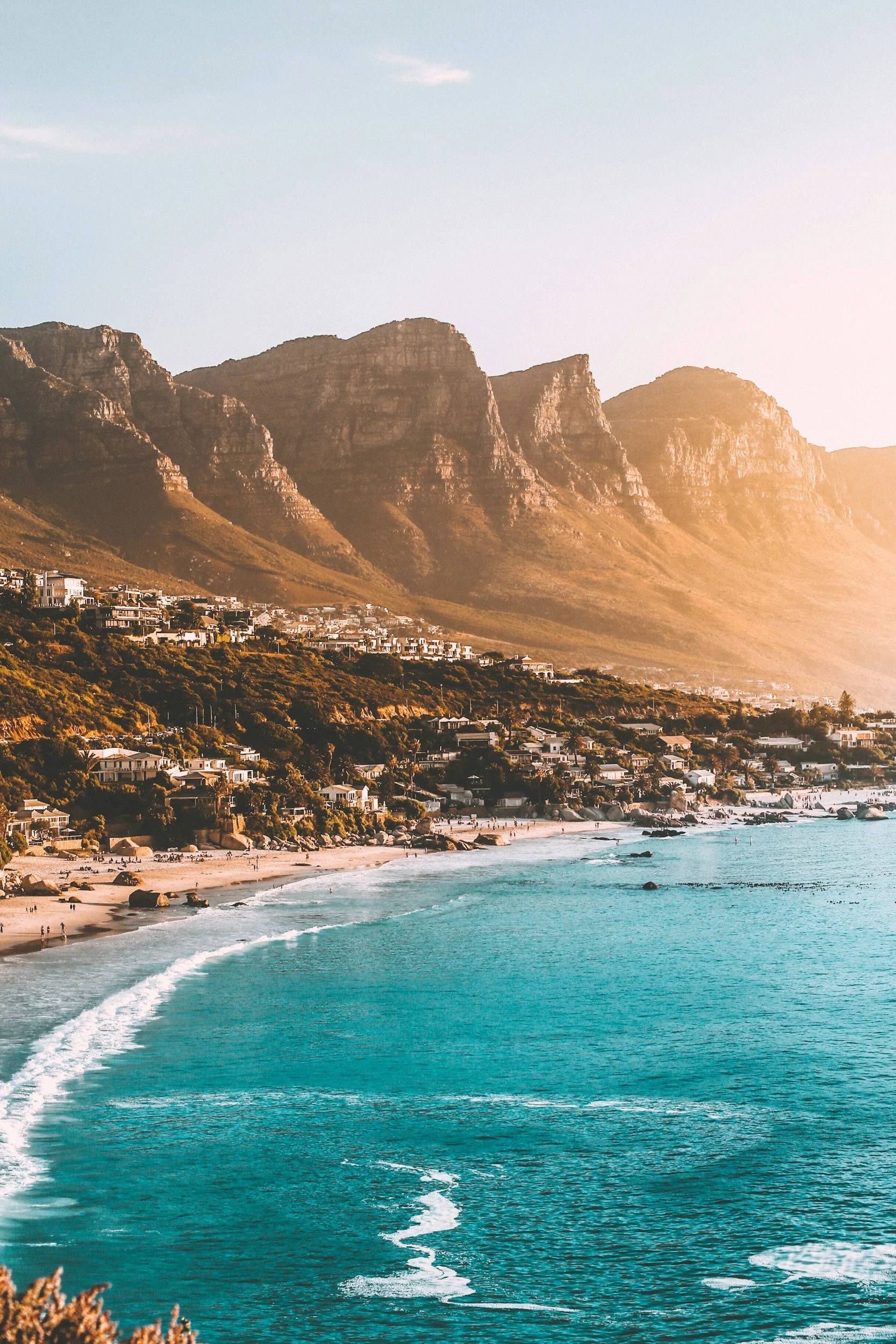 A scenic view of a beach with turquoise water and waves, a sandy shoreline, and houses along the coast, with large mountains in the background during sunset.