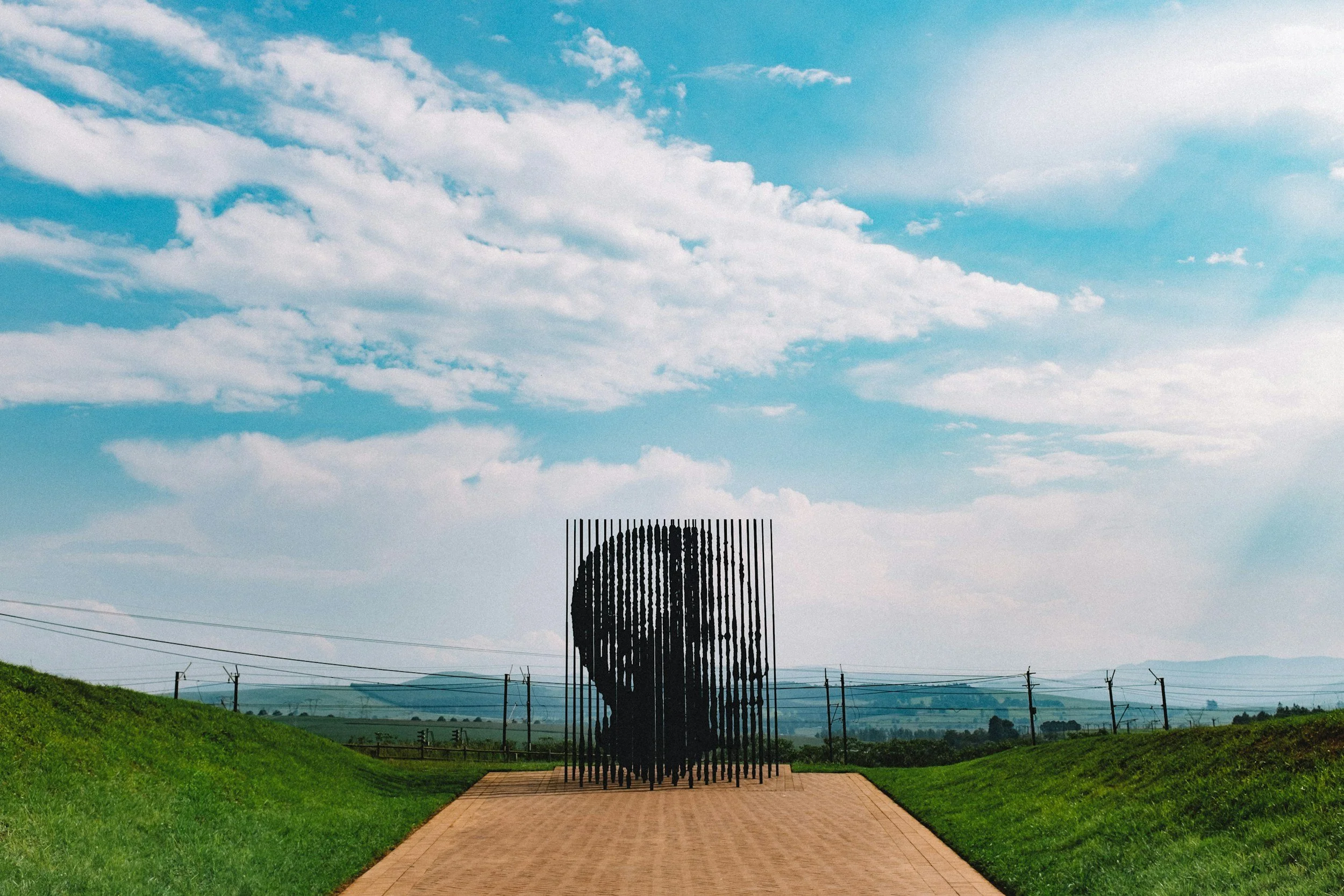 Outdoor sculpture of a human face created from vertical metal rods, set in a grassy landscape under a blue sky with clouds.