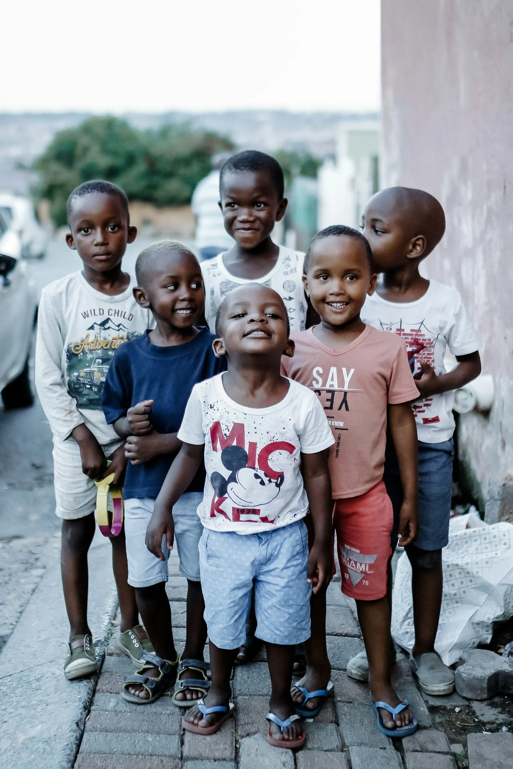 Group of seven smiling children standing on a sidewalk next to a wall in an urban area.