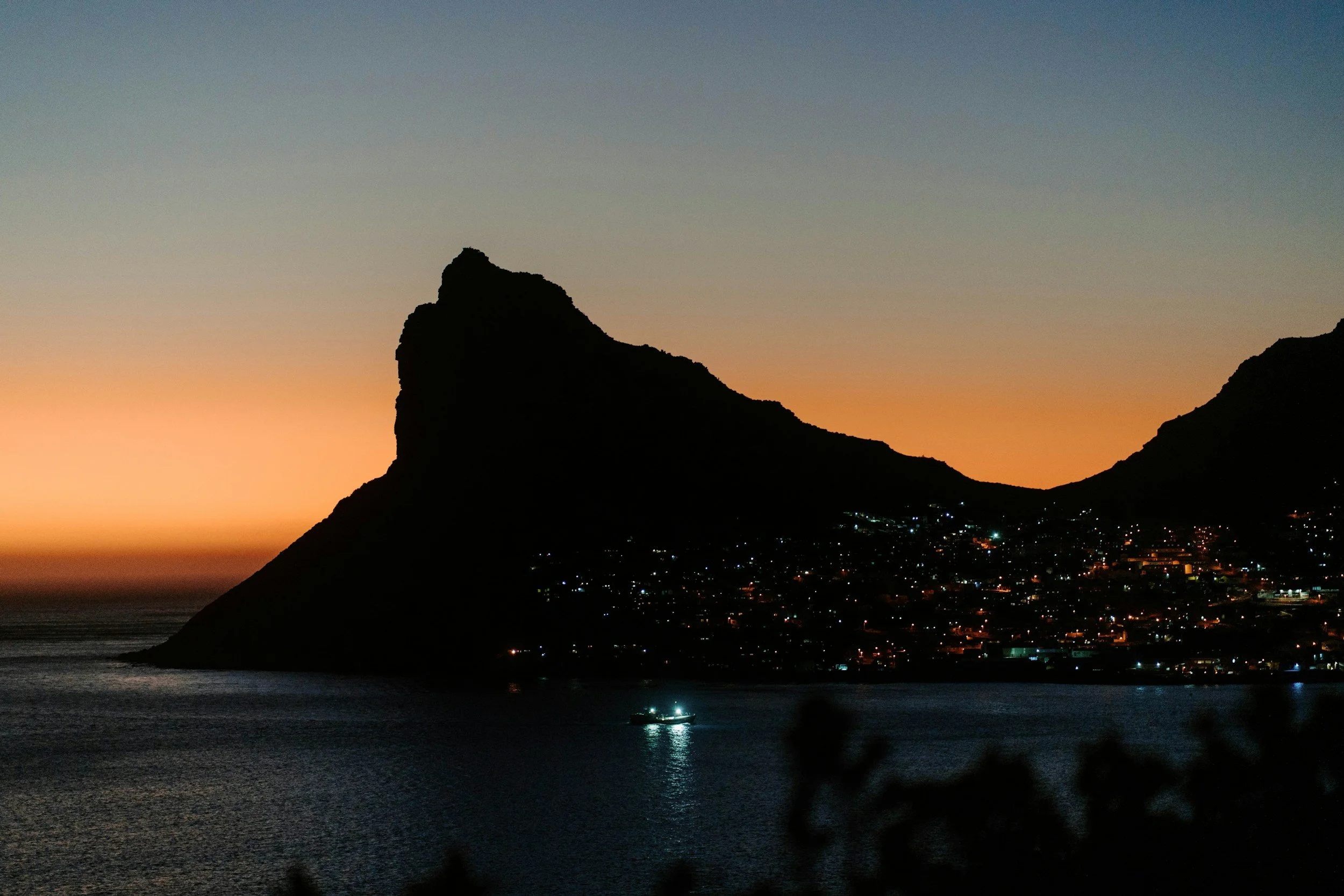 Silhouette of a mountain by the ocean at sunset, with a small boat on the water and a city with lights in the distance.