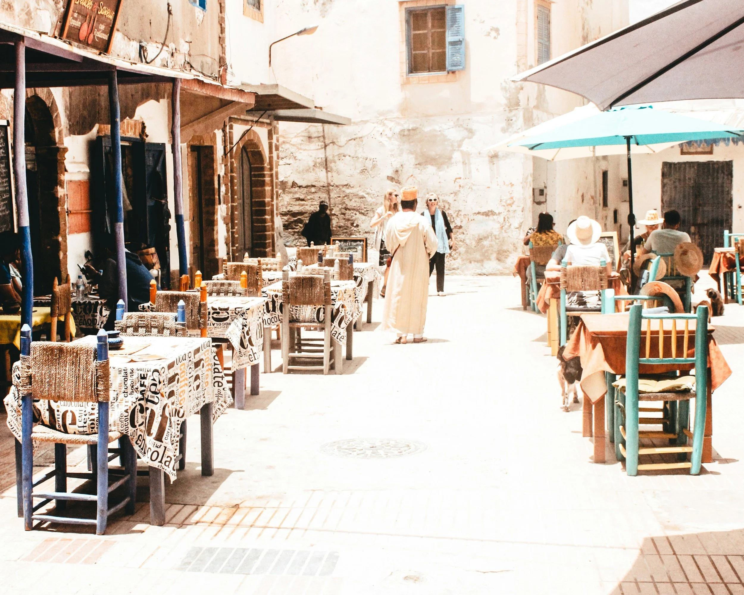 Outdoor café with tables and chairs, some under umbrellas, in a sunny European street with a few people sitting and walking.