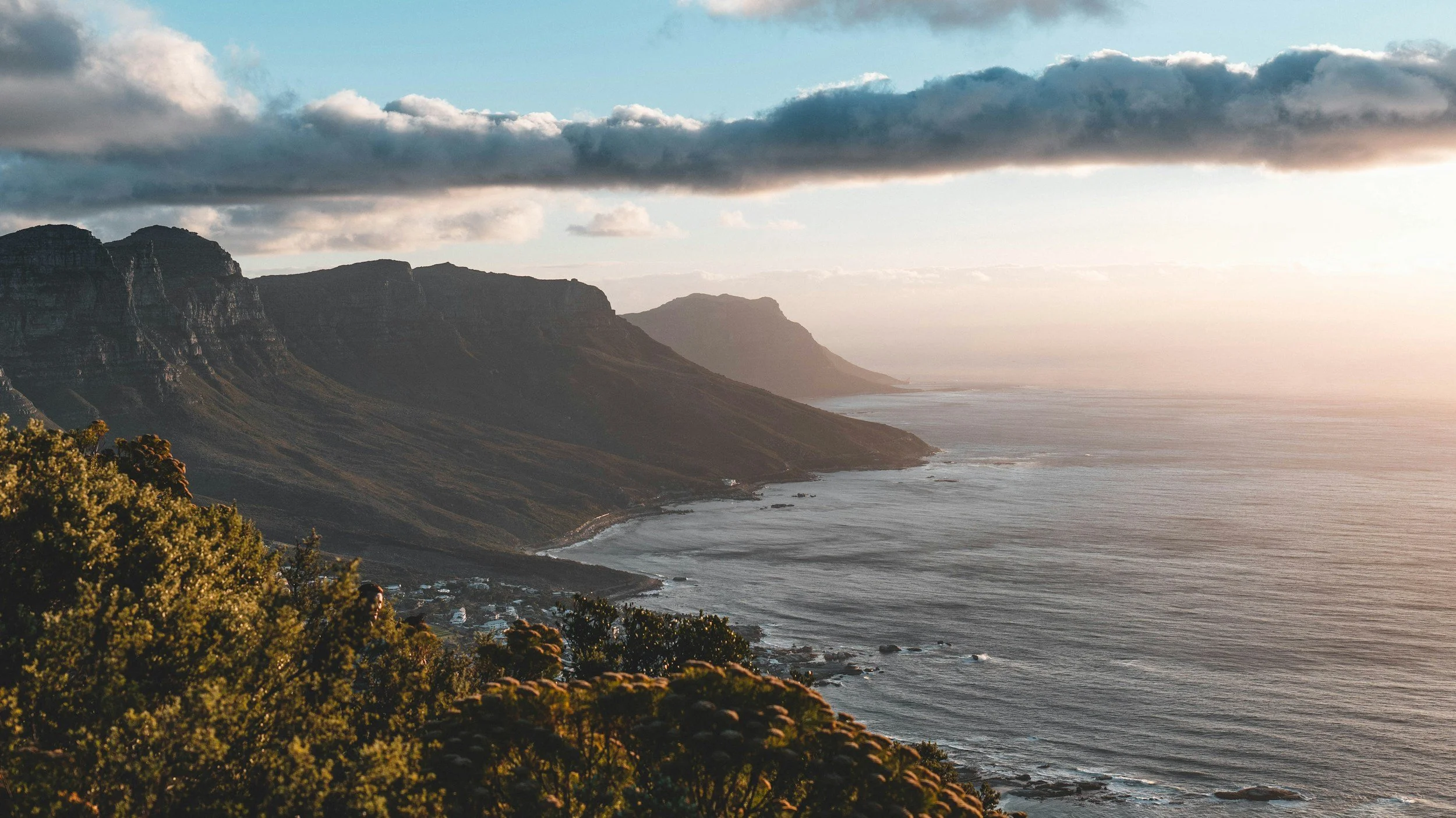 Sunset over rugged coastal cliffs with ocean waves and greenery in the foreground.