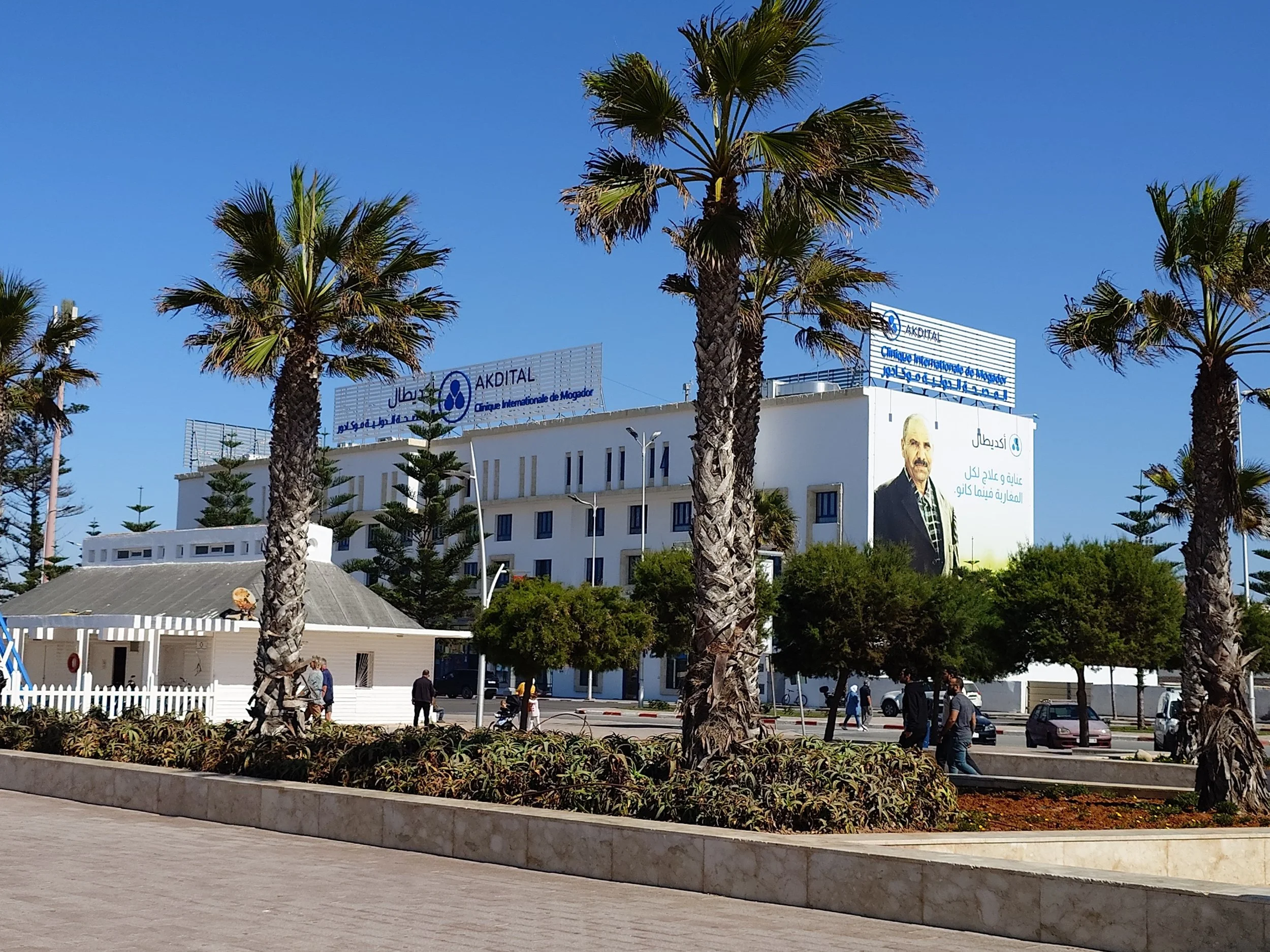 A white multi-story building with signs in Arabic and French, surrounded by tall palm trees and some smaller trees, on a bright sunny day with a clear blue sky.