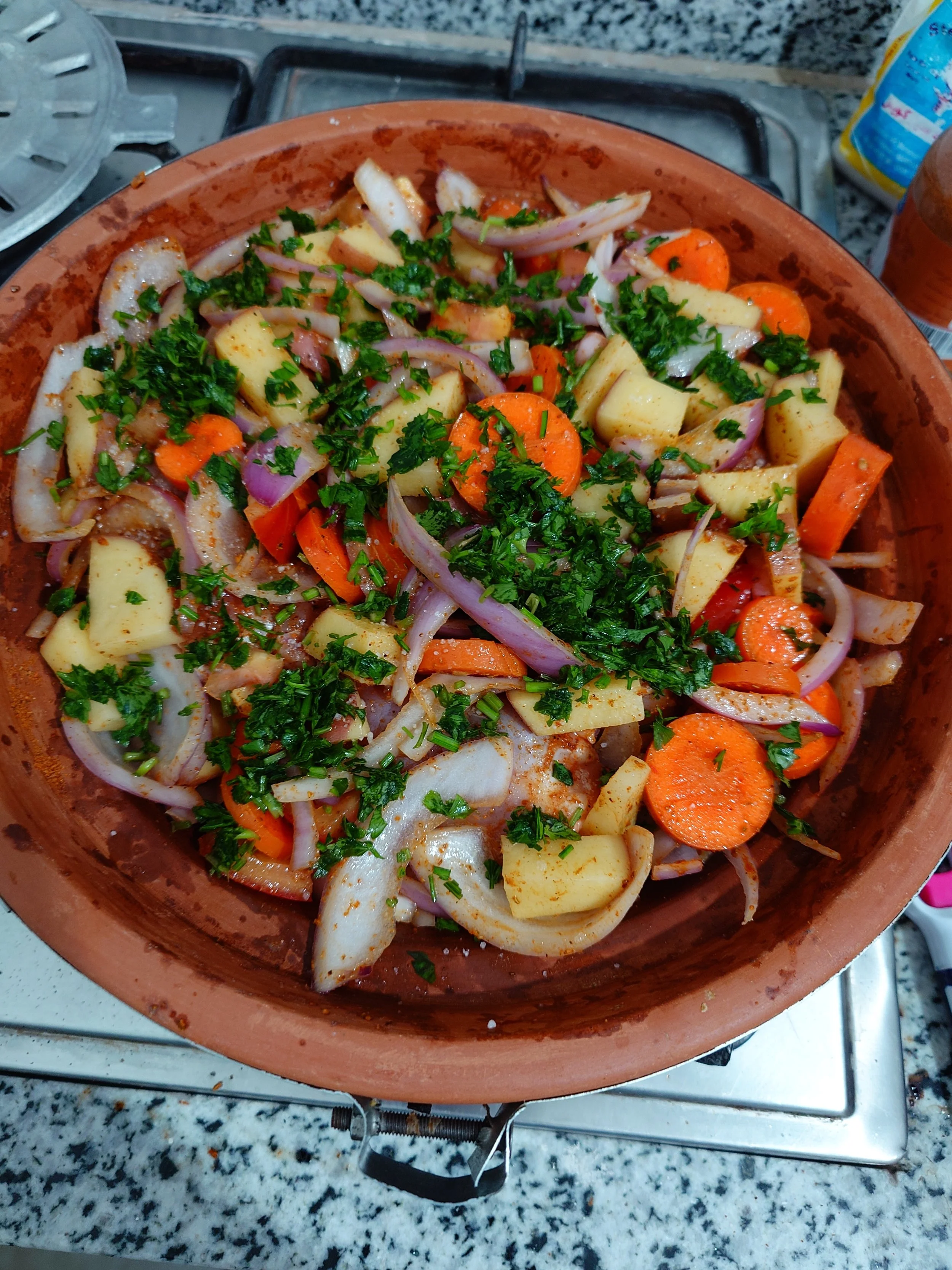 A terracotta bowl filled with chopped potatoes, carrots, onions, and herbs on a stove top.