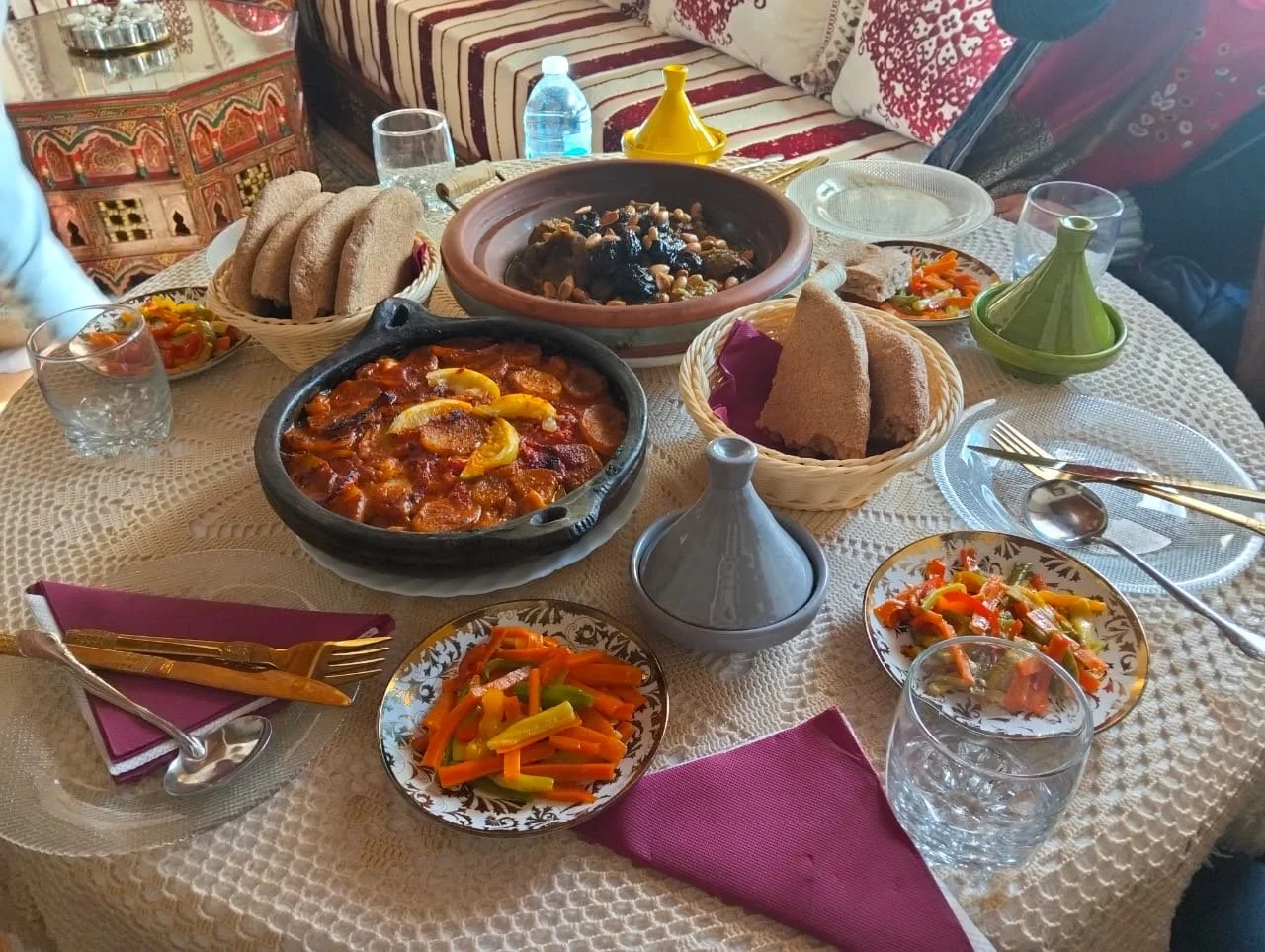A Moroccan-style dinner table set with bread, vegetable stew, tagine dishes, colorful vegetable salads, and drinking glasses on a lace tablecloth.