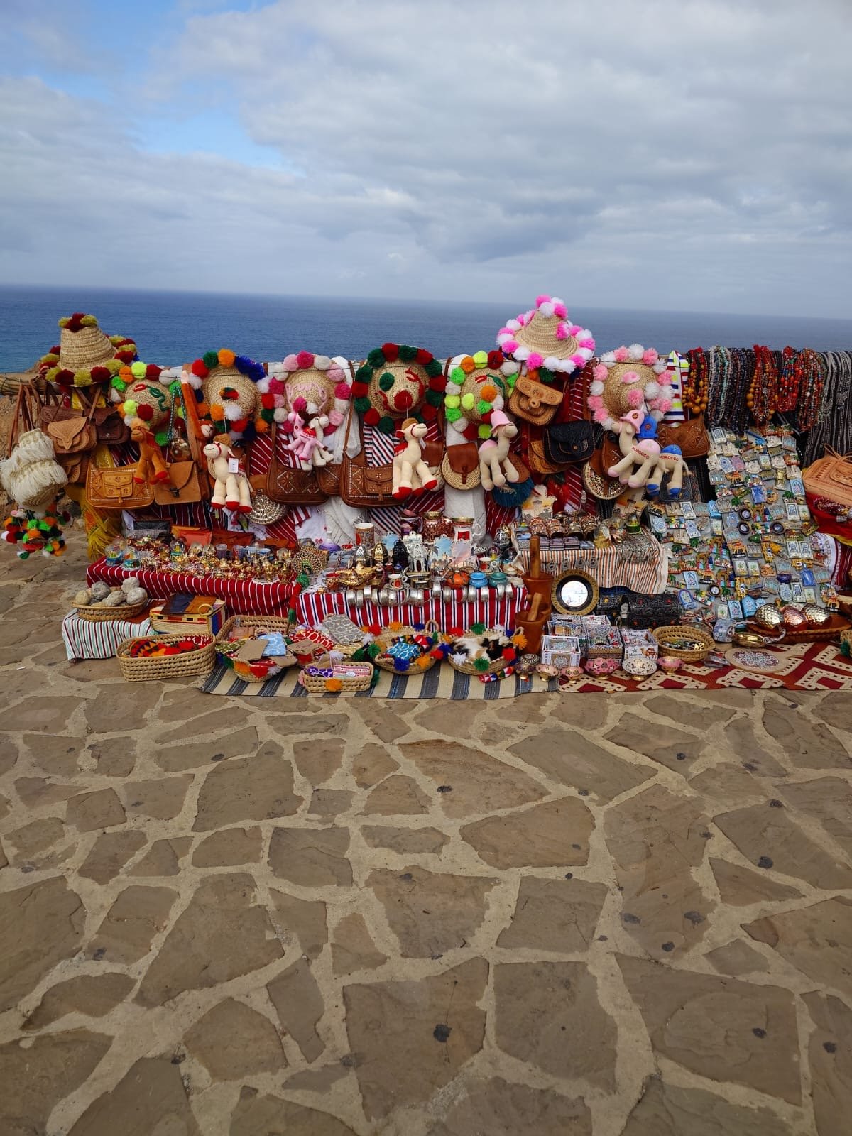 Colorful outdoor market stall with handmade crafts, jewelry, and toys, set against a view of the ocean and cloudy sky.