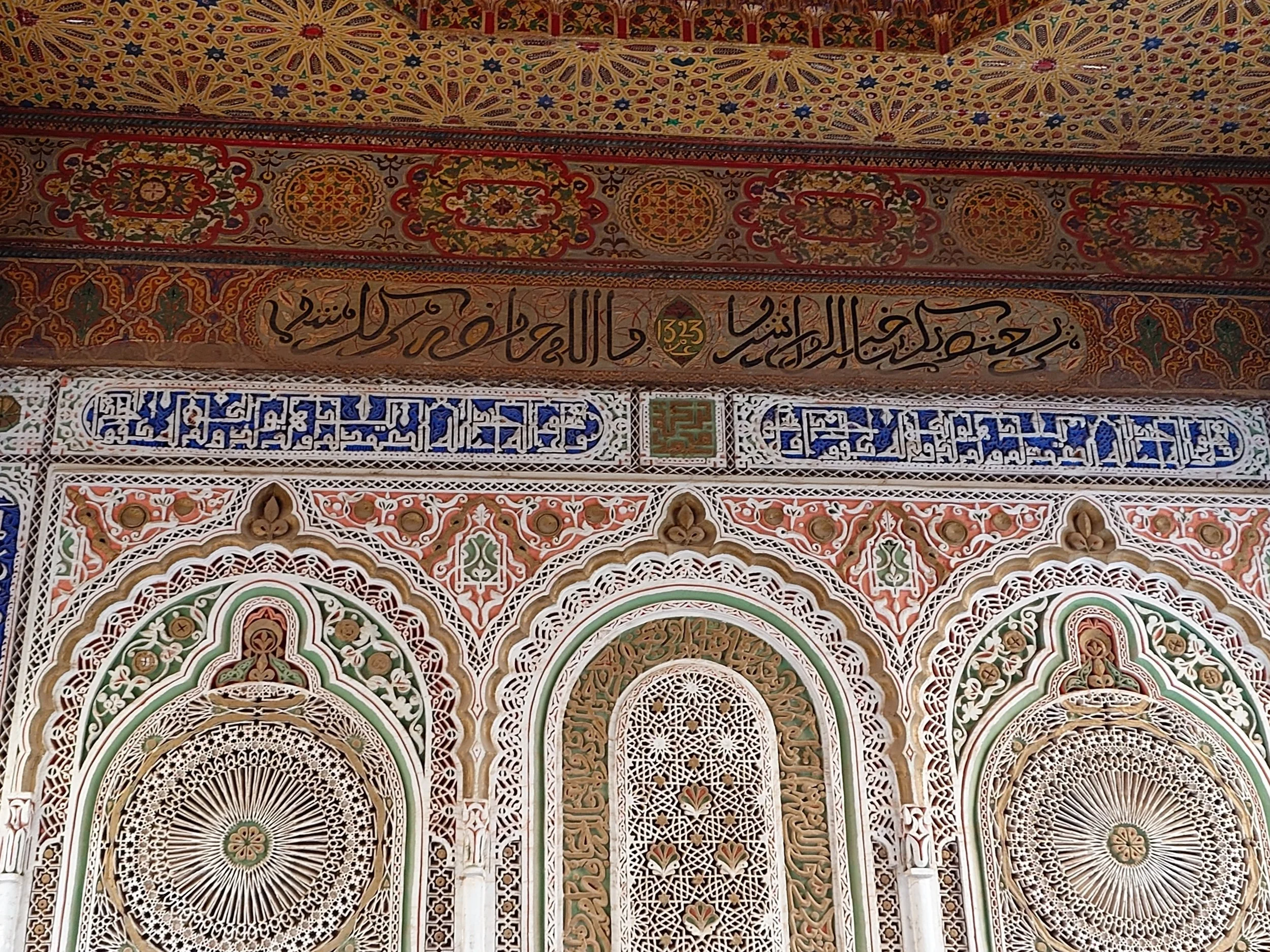 Intricate Islamic architectural details with Arabic calligraphy, geometric and floral patterns in colors of white, green, red, gold, and blue, likely part of a monument or mosque interior.