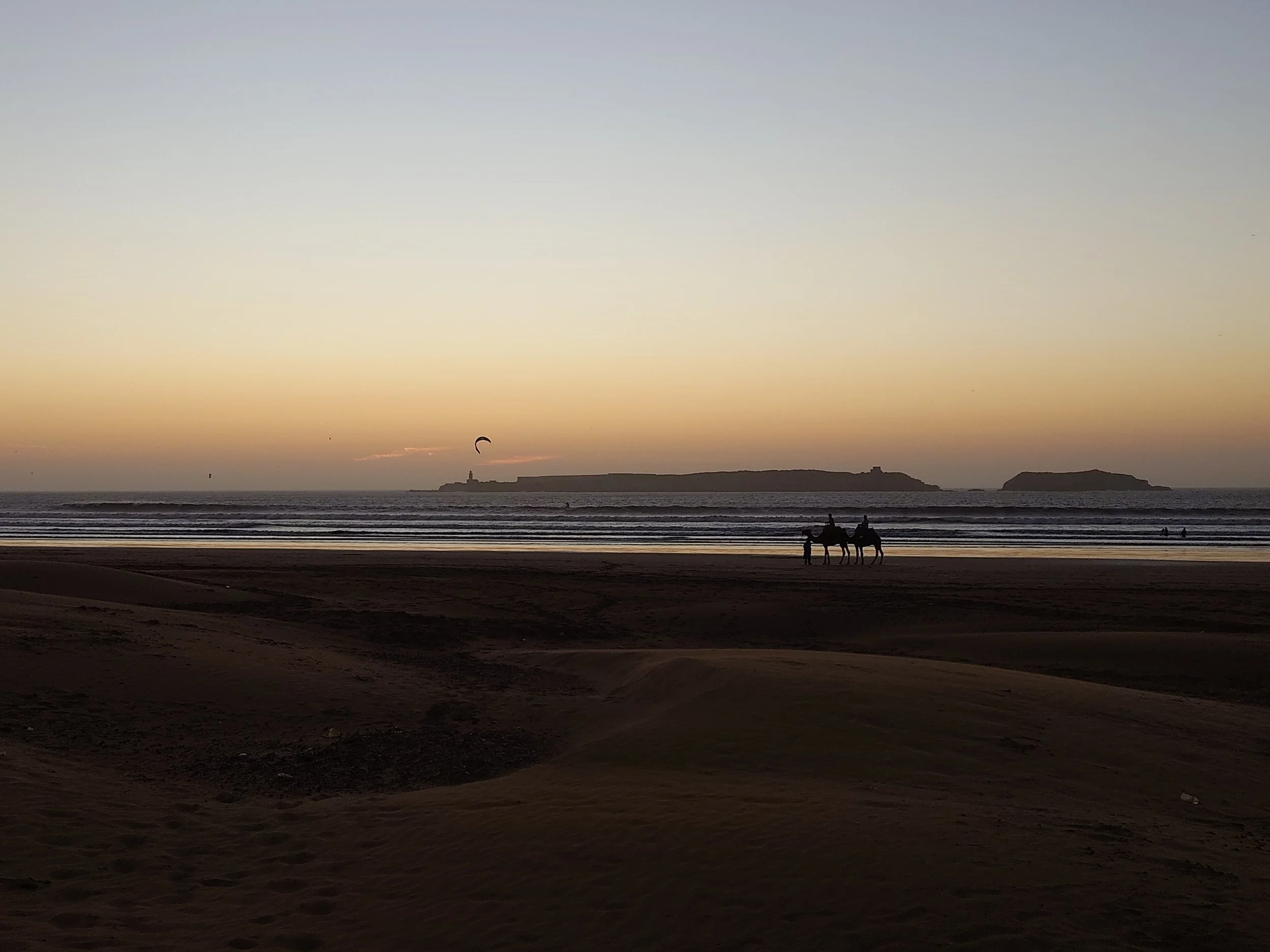 Silhouettes of two people riding horses along a beach at sunset, with a lighthouse on a distant island and a kite surfer in the sky.