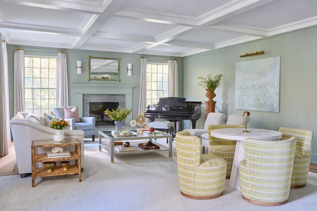 A spacious, well-lit living room with large windows, a white sofa and armchair, a gray armchair near the fireplace, a black grand piano, a round dining table with four patterned chairs, a coffee table with decorative items, and potted plants, with so