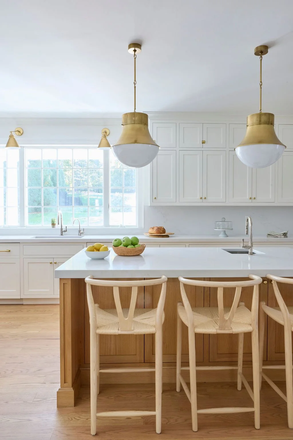 Bright kitchen with white cabinets, large window, and wooden island. Three hanging pendant lights with gold accents. Bowls of green apples and yellow lemons on the island. Natural light streaming in.
