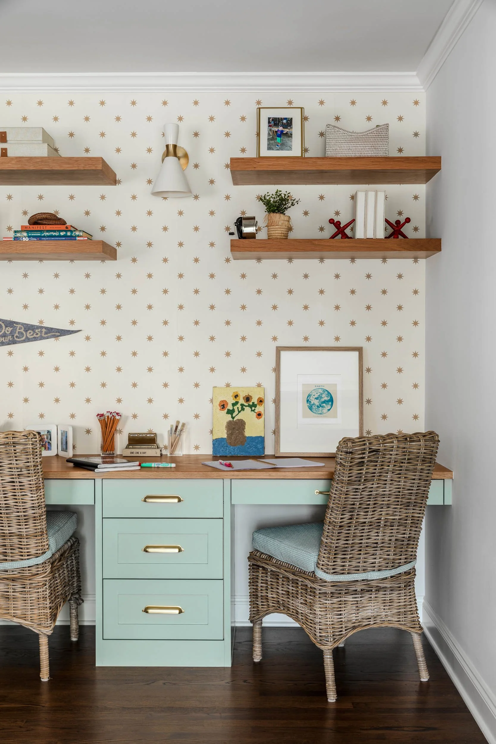 A home office or study area featuring a mint green desk with gold handles, surrounded by wicker chairs, with wall-mounted wooden shelves holding books, plants, and decorative objects, against a wall with star-patterned wallpaper.