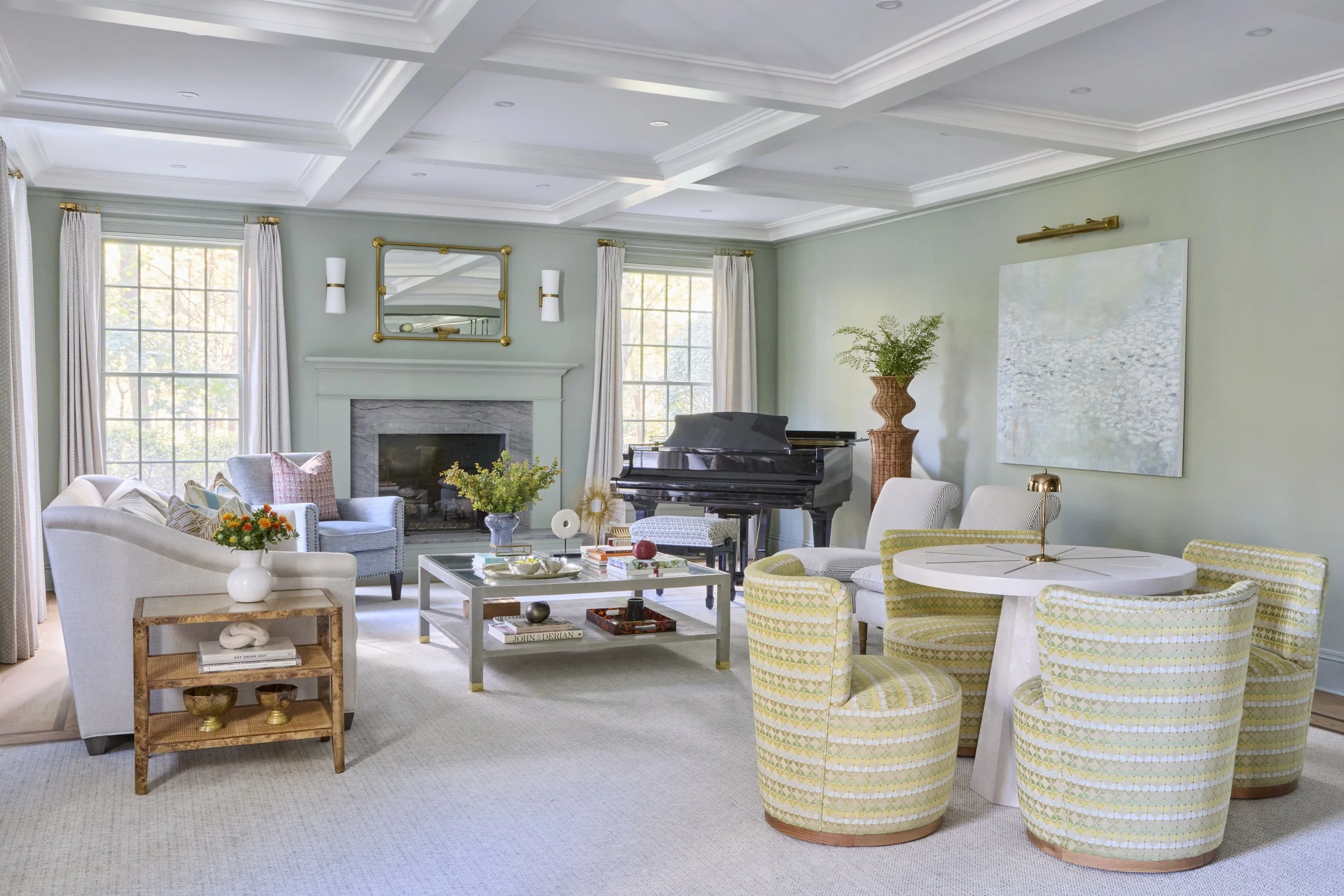A spacious, well-lit living room with large windows, a white sofa and armchair, a gray armchair near the fireplace, a black grand piano, a round dining table with four patterned chairs, a coffee table with decorative items, and potted plants, with so