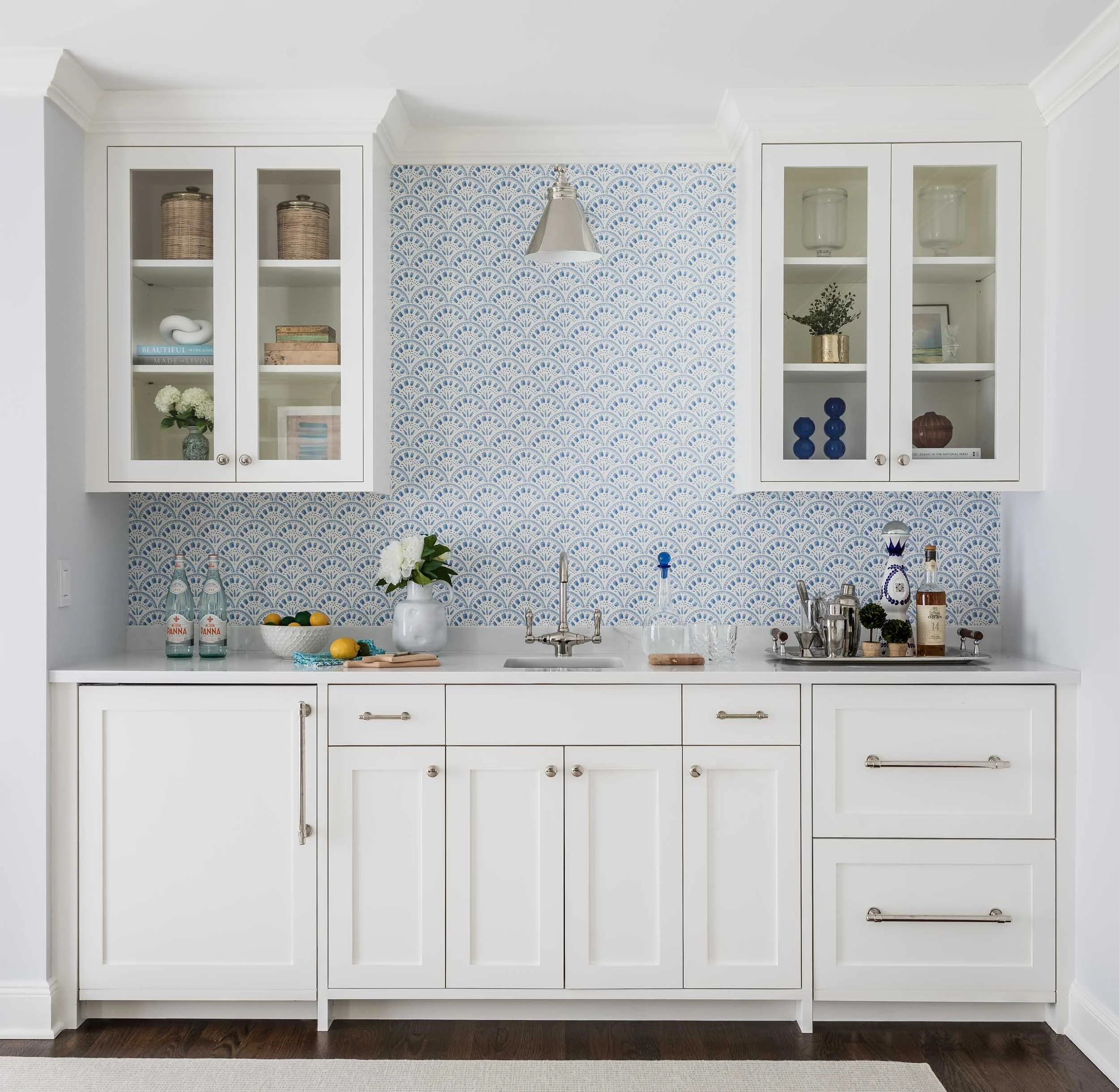 A white kitchen cabinet with glass doors on top, decorated with woven baskets, books, a plant, and bottles inside. The wall behind has blue and white patterned wallpaper, and the countertop has flowers, bottles, glasses, and kitchen utensils.
