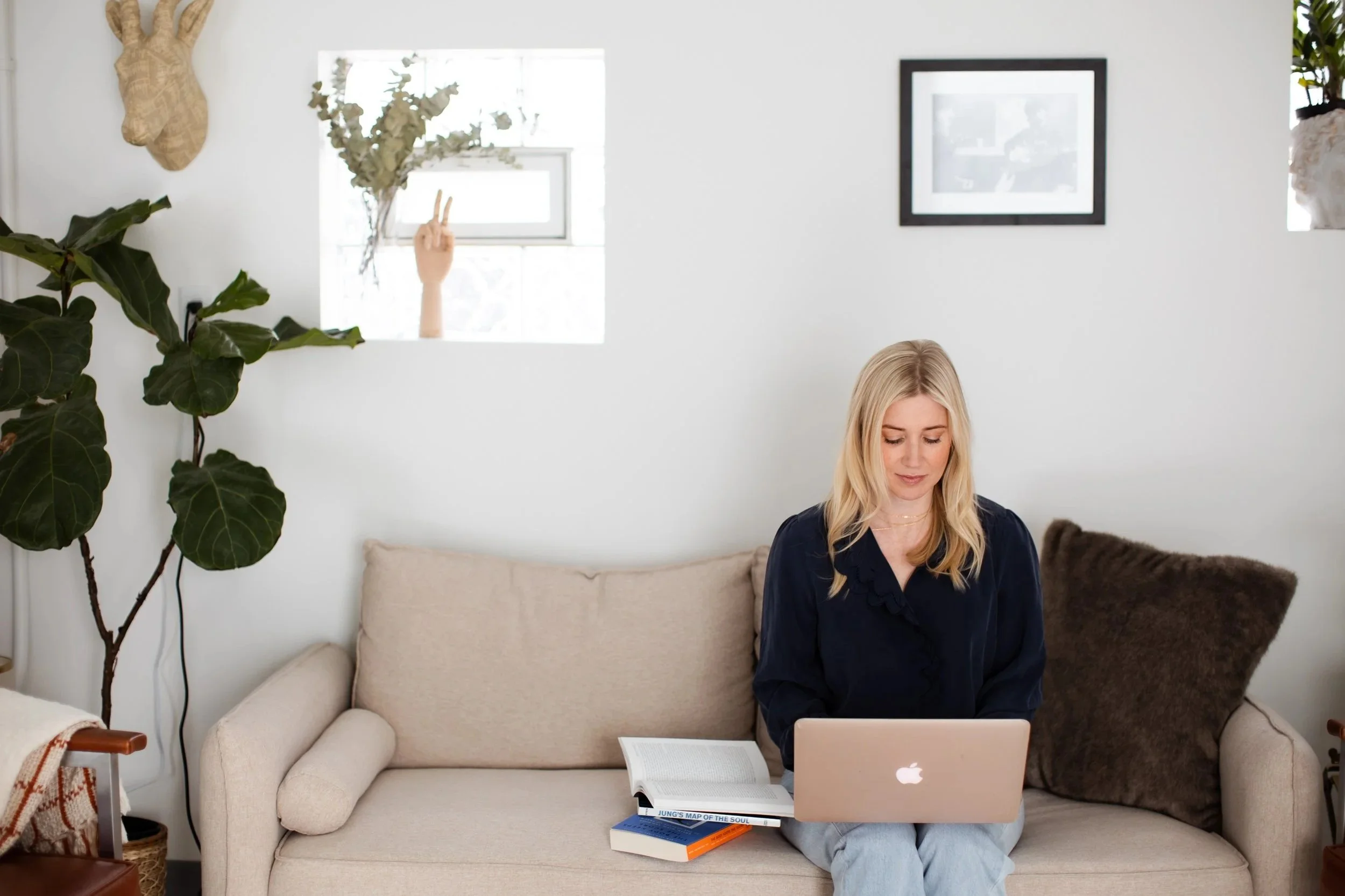A woman sitting on a beige couch working on a laptop with books and an open book on her lap, in a room with a large green leafy plant, framed pictures, and a wall-mounted decorative object.