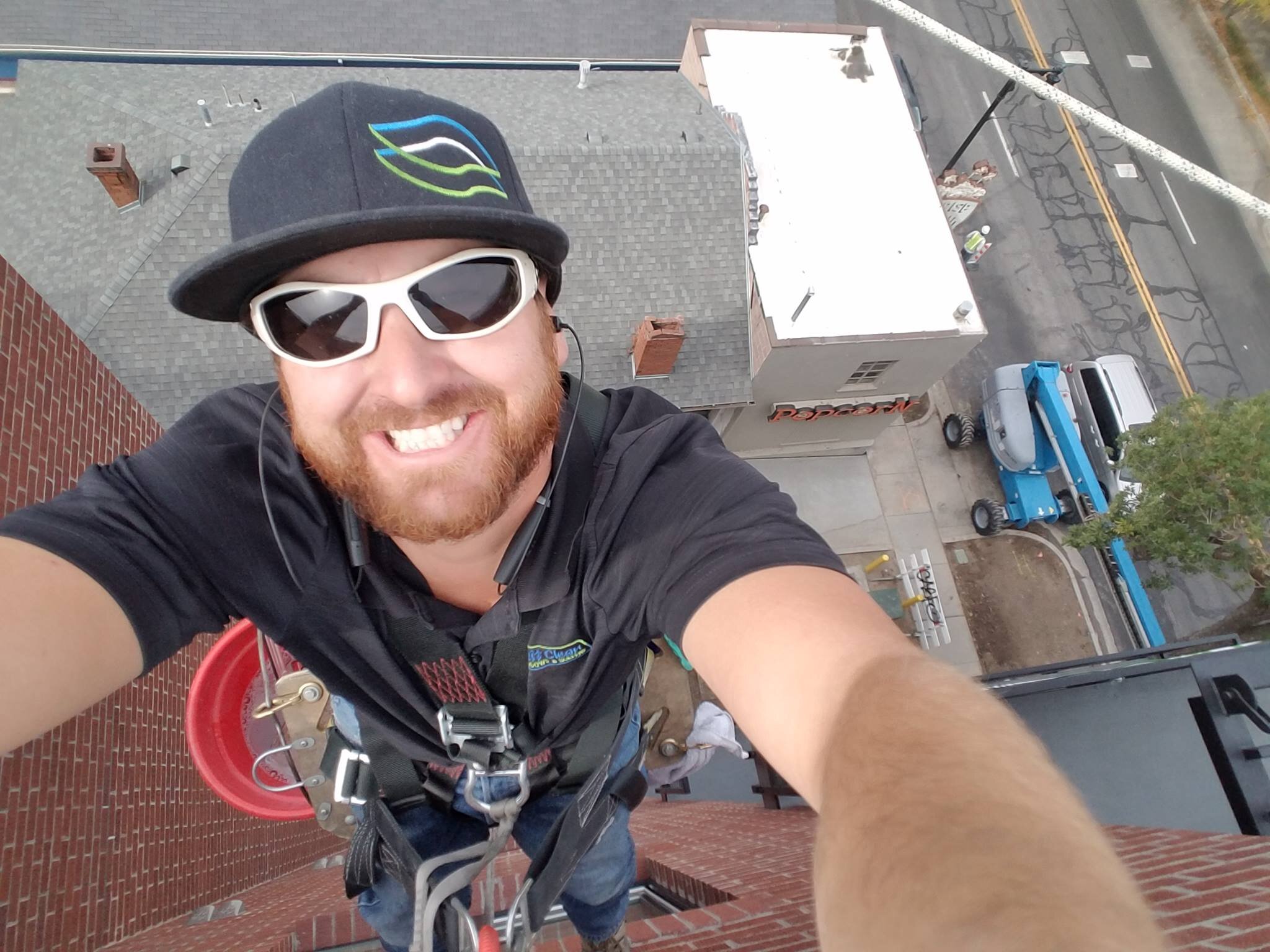 A window washer in Salt lake city Utah near mewith a beard and sunglasses taking a selfie from a high rooftop during daytime, with a city street and buildings visible below.