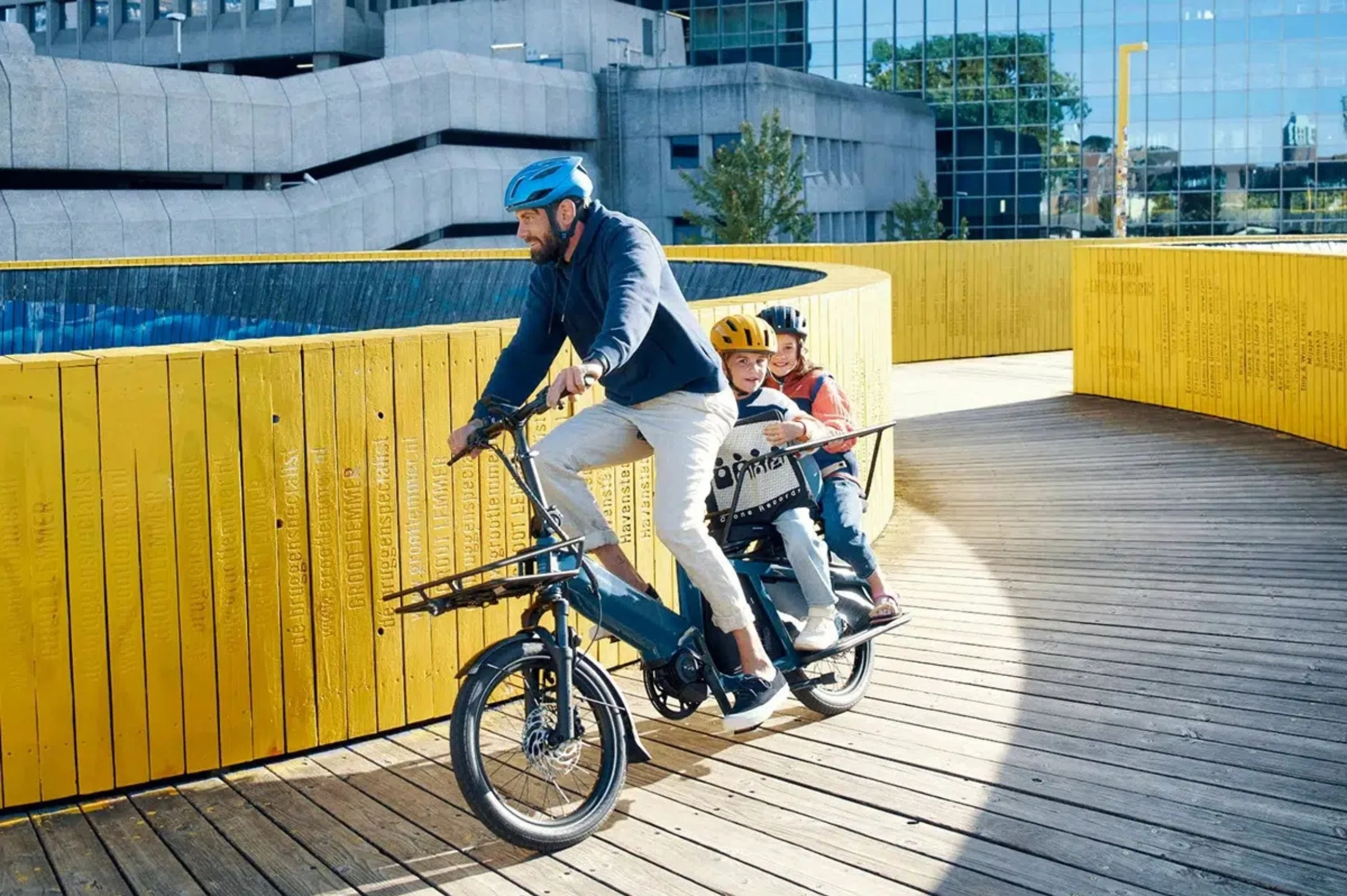 A man riding a cargo bike with two children in helmets on a wooden walkway.