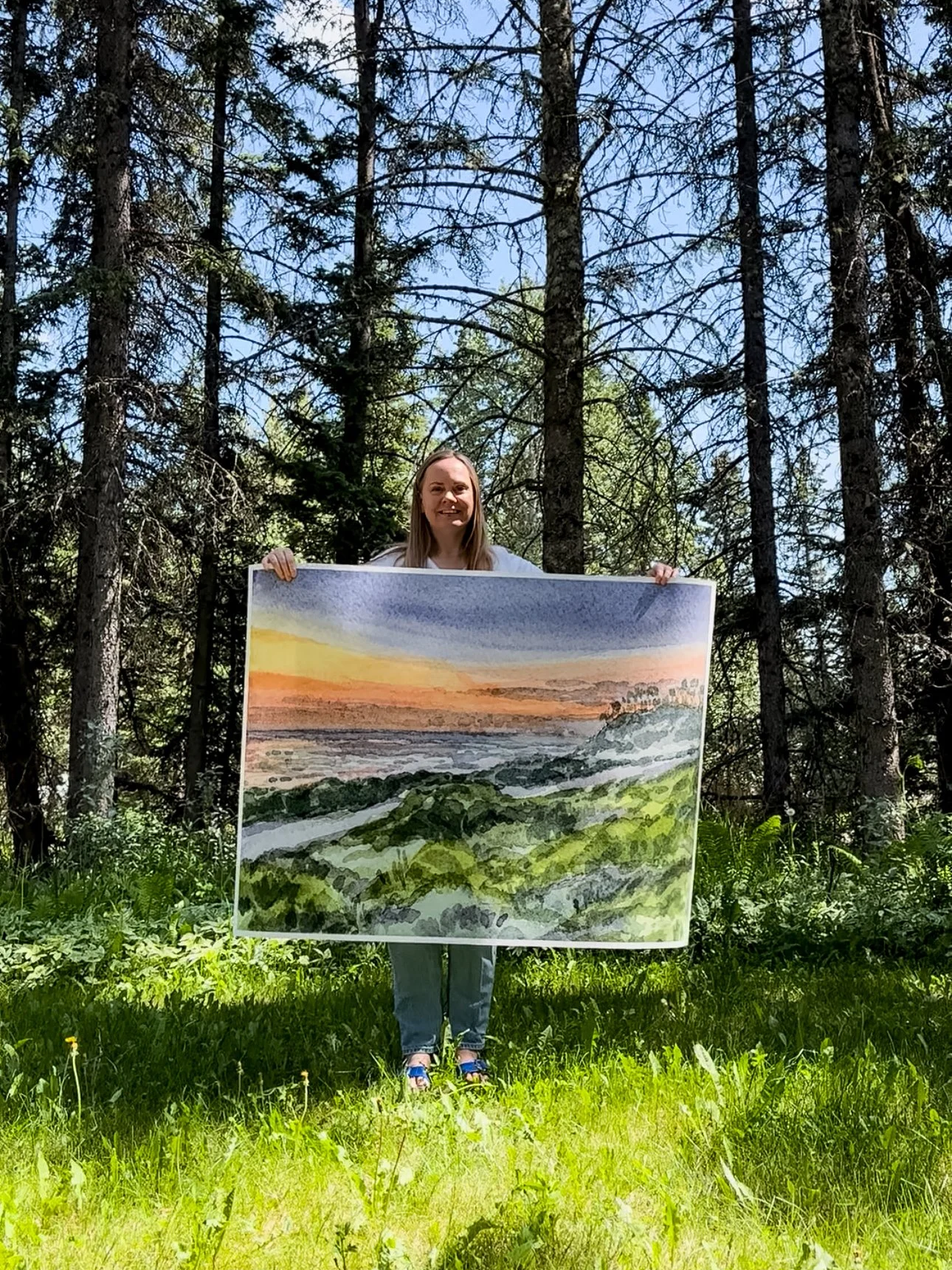 Artist, Rebecca Sobbi holding a large watercolor print of a vibrant beach sunset from a bike trail in Encinitas, CA.