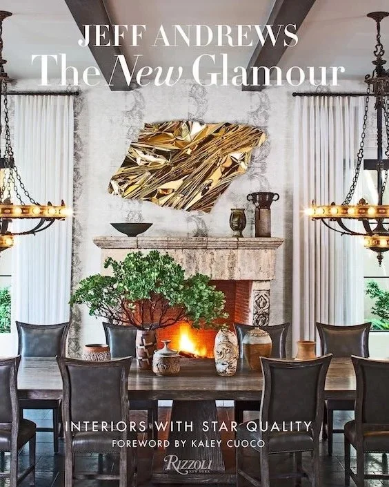 Jeff Andrews' book cover for The New Glamour: Interiors with Star Quality with Foreword by Kayley Cuoco published by Rizzoli with a large dining room table and chairs in front of a warm fire with a rich marble mantle & striking gold textured art hung