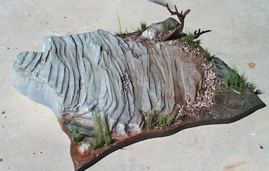 I added model railroad gravel to the base of the cliff and little bits in crevasses. I watered down Elmers glue and sprayed it over the gravel to lock it in place. I added some railroad grass (very fine green threads that you cut to size) to the base as well as a dead tree that I recently discovered in a box.