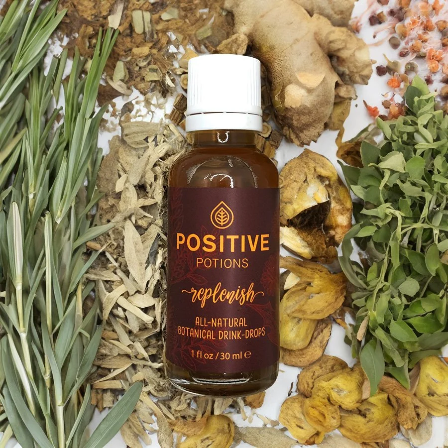 A brown bottle labelled "Positive Potions Replenish All-Natural Botanical Drink-Drops" surrounded by dried herbs, roots, and flowers on a white surface.