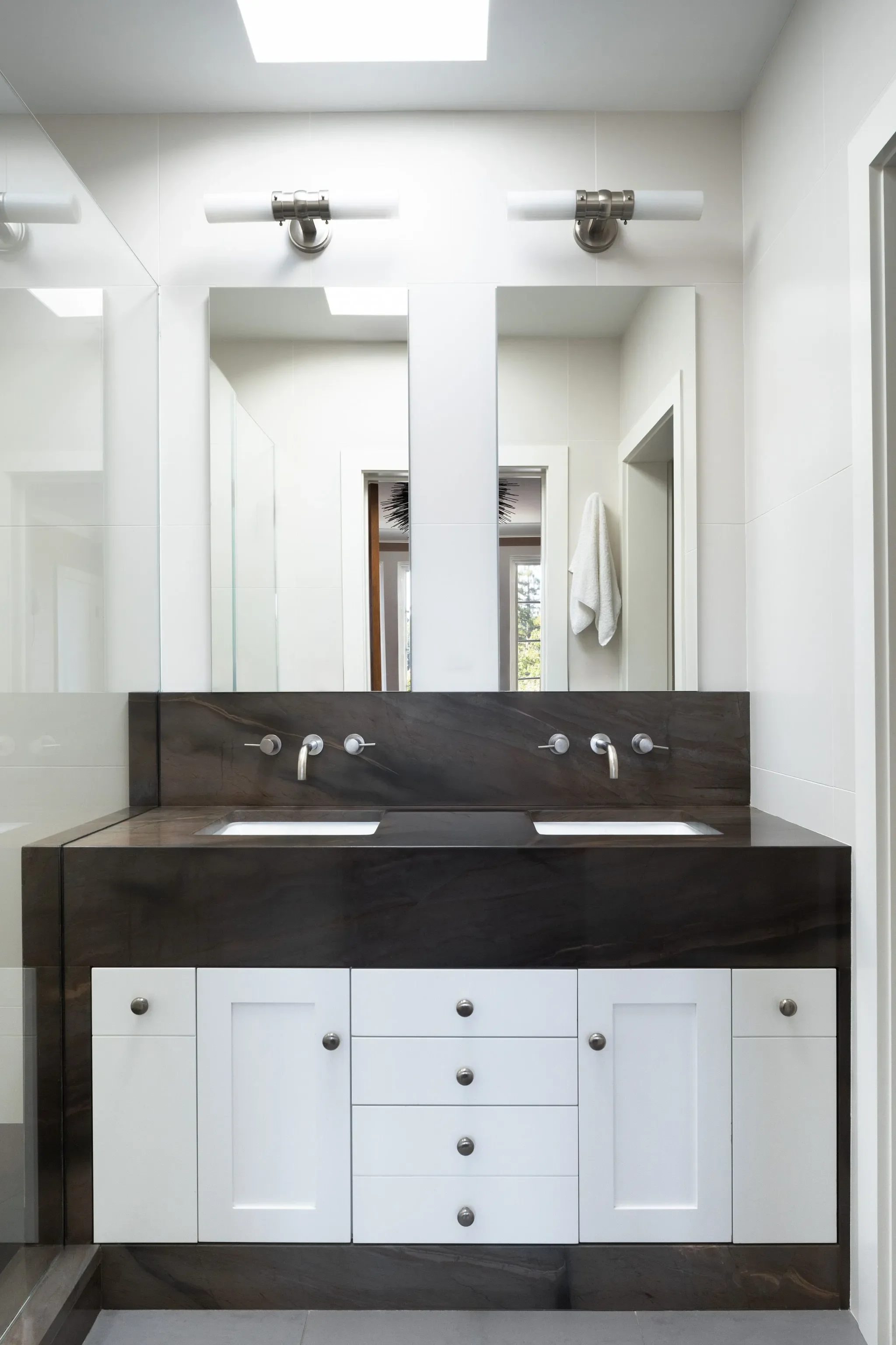 Modern bathroom featuring a large mirror, two ceiling lights above, white walls, a white towel hanging on the right side, and a dark countertop with a sink and multiple drawers.