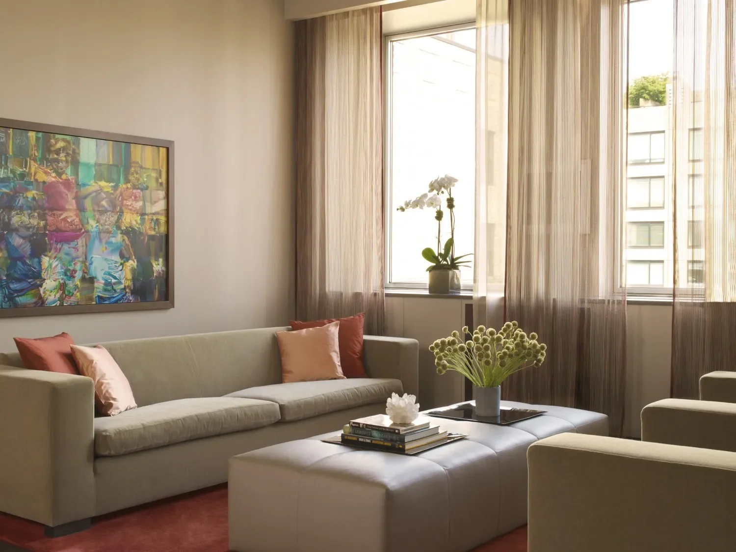 A beige living room with a cream sofa with pink cushions, a white ottoman with a stack of books and a vase of flowers, beige curtains, a large window with blinds, a potted plant on the windowsill, and an abstract painting on the wall.