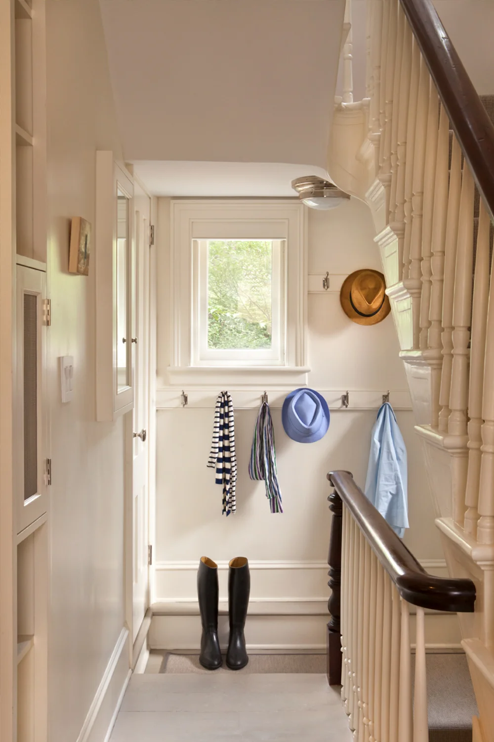 A small entryway with hooks on the wall holding scarves and hats, a pair of black rain boots on the floor, and a window allowing natural light into the area.