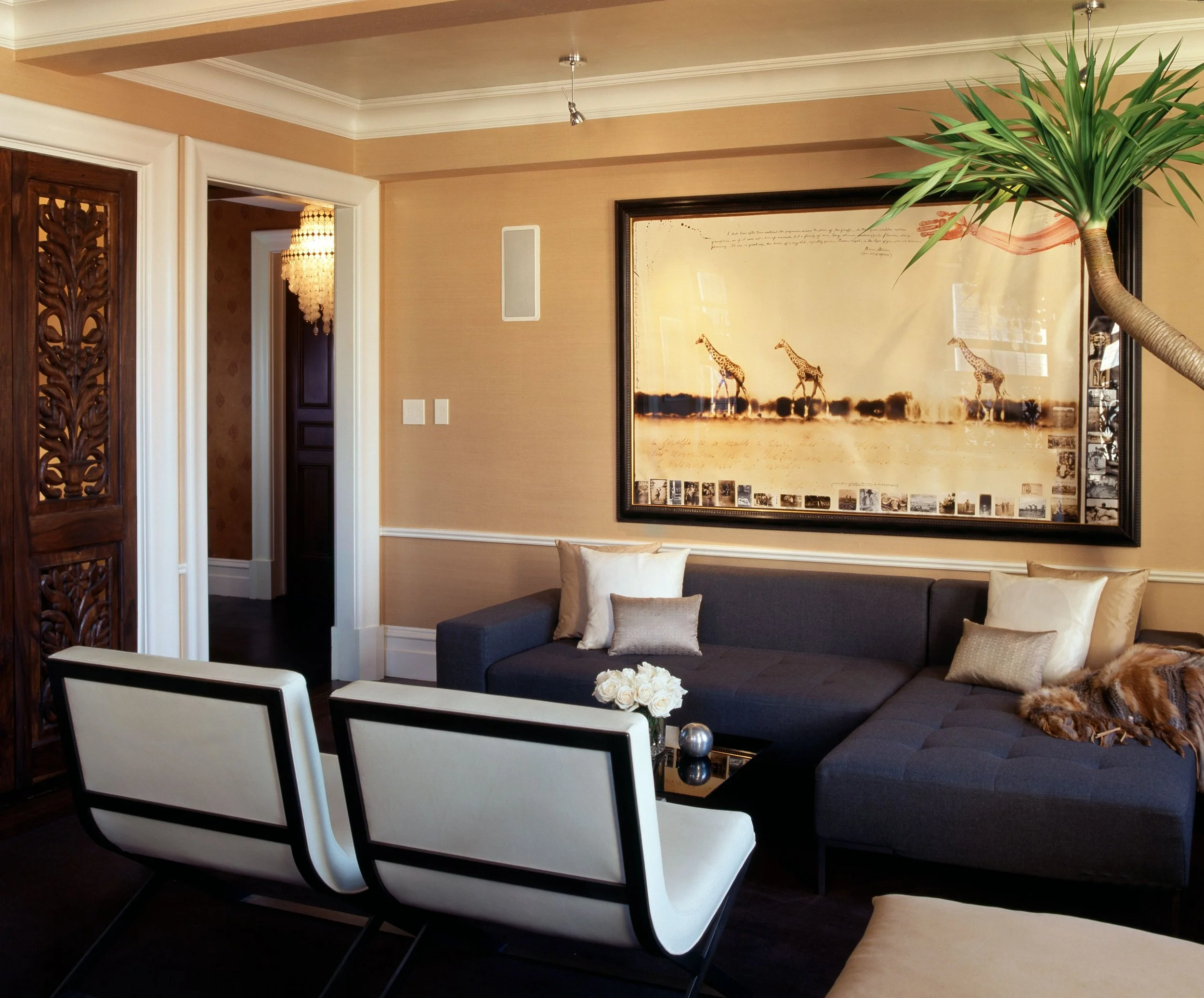 Warm living room with navy sectional, modern white chairs, and large framed giraffe artwork on gold walls
